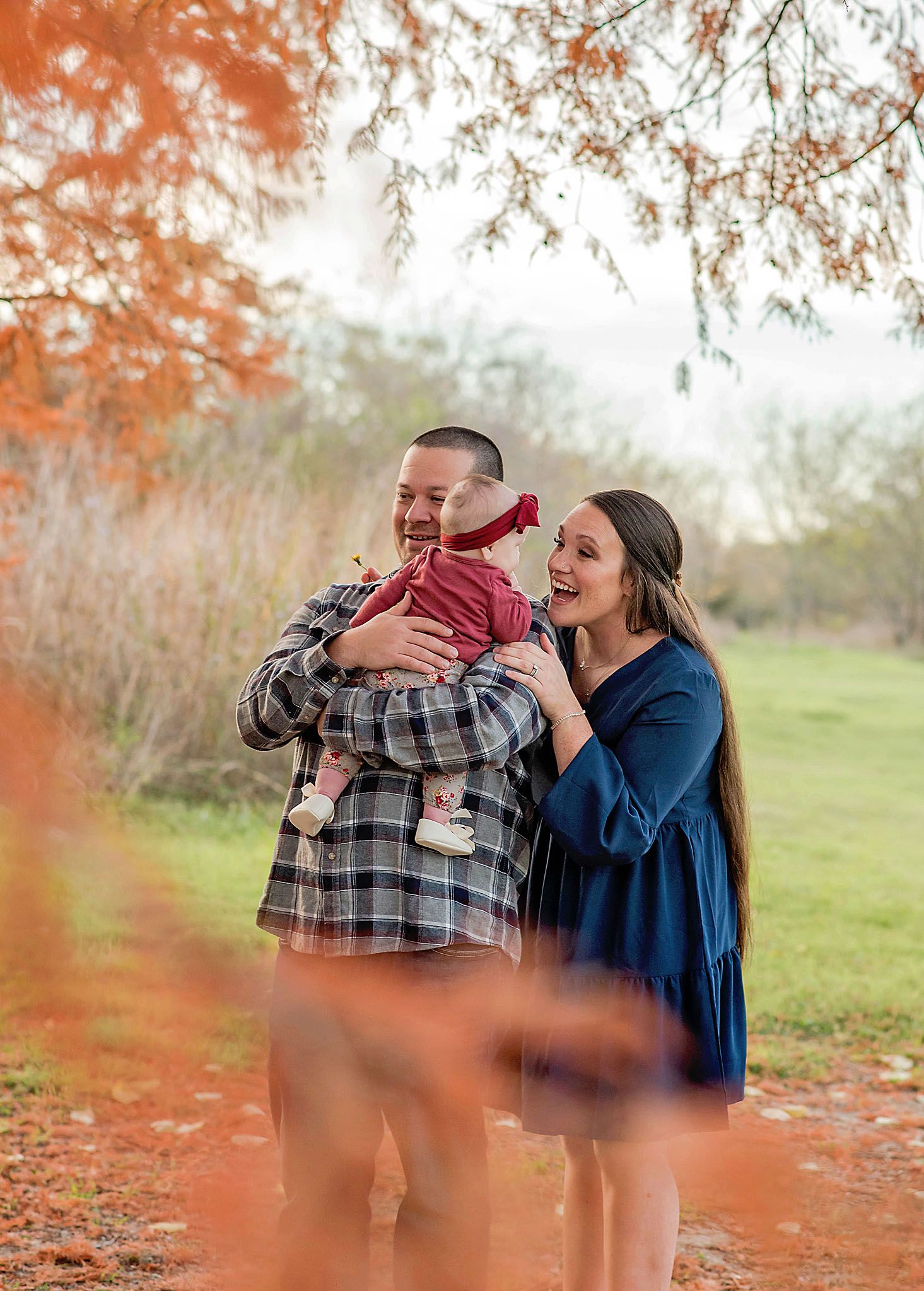 A couple stands together with a baby in autumn, framed by orange fall foliage in a peaceful outdoor setting.