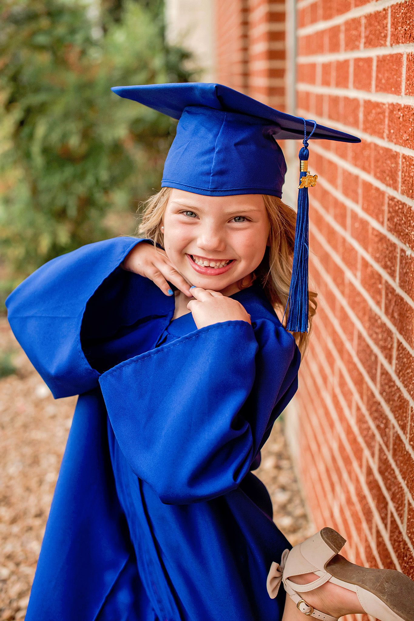 A smiling young girl in a blue graduation cap and gown poses proudly against a brick wall.