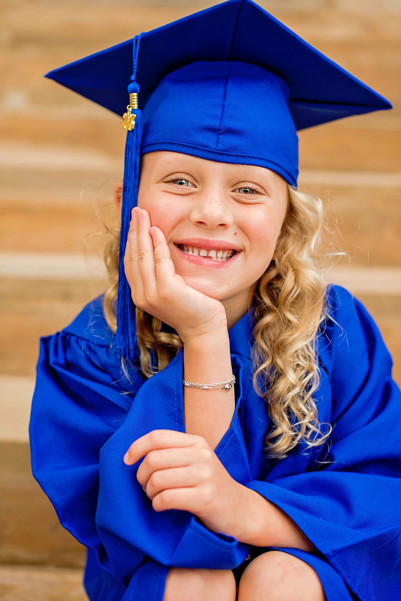 Young girl in blue graduation cap and gown smiling proudly, resting chin on hand for kindergarten graduation photo.