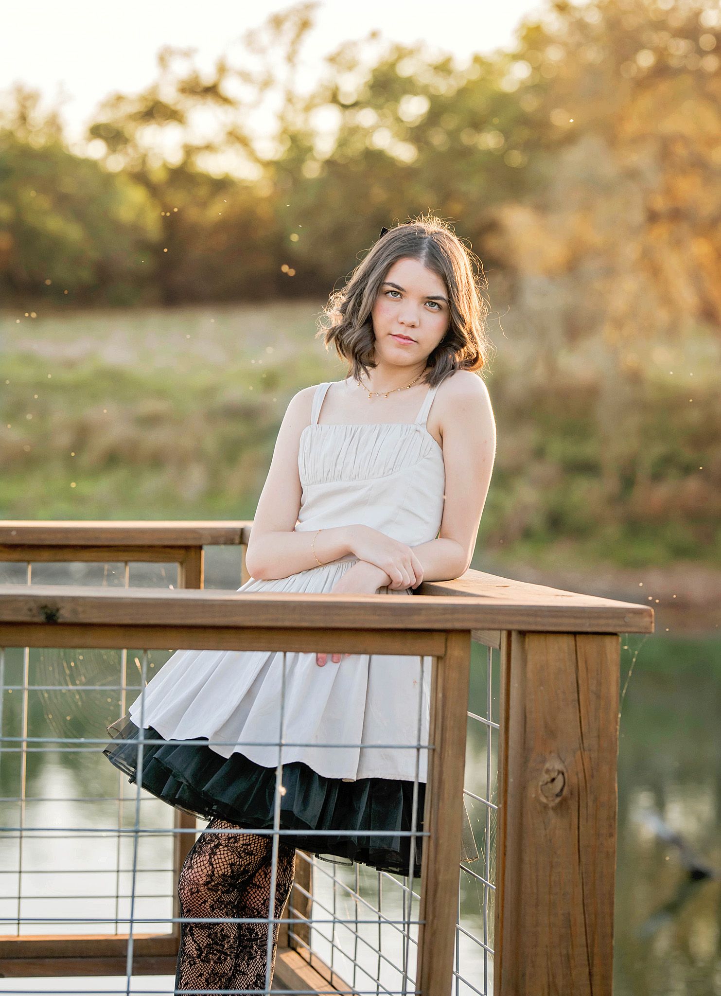 A teenage girl in a white top leans on a wooden railing outdoors during golden hour near a pond.