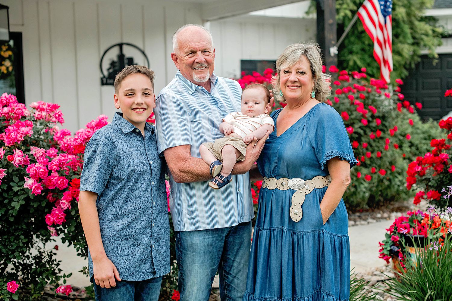 Grandparents with grandson and baby pose by colorful roses and American flag.