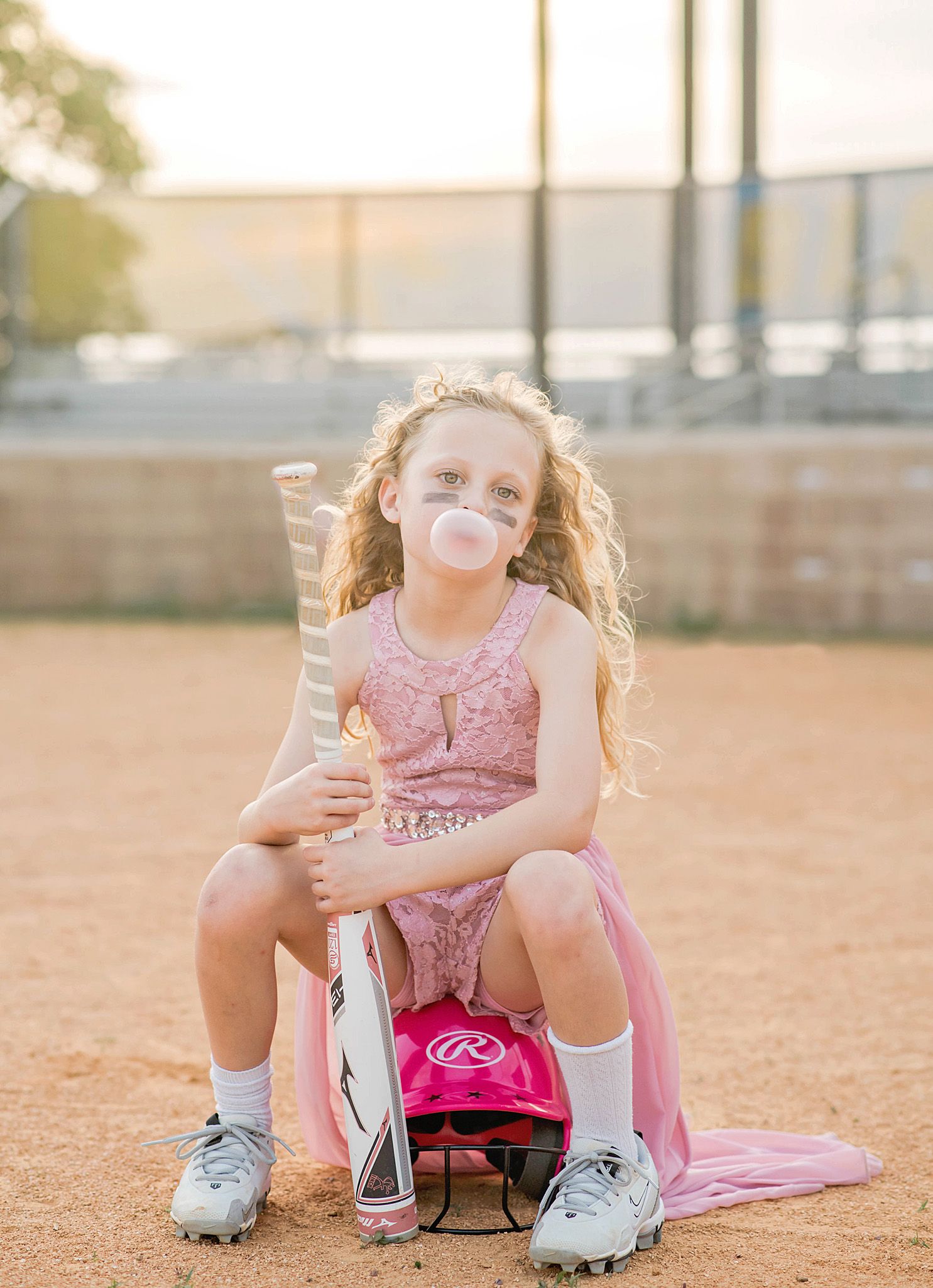 Young girl in pink softball uniform blowing bubble gum, holding bat on dirt field with pink helmet.