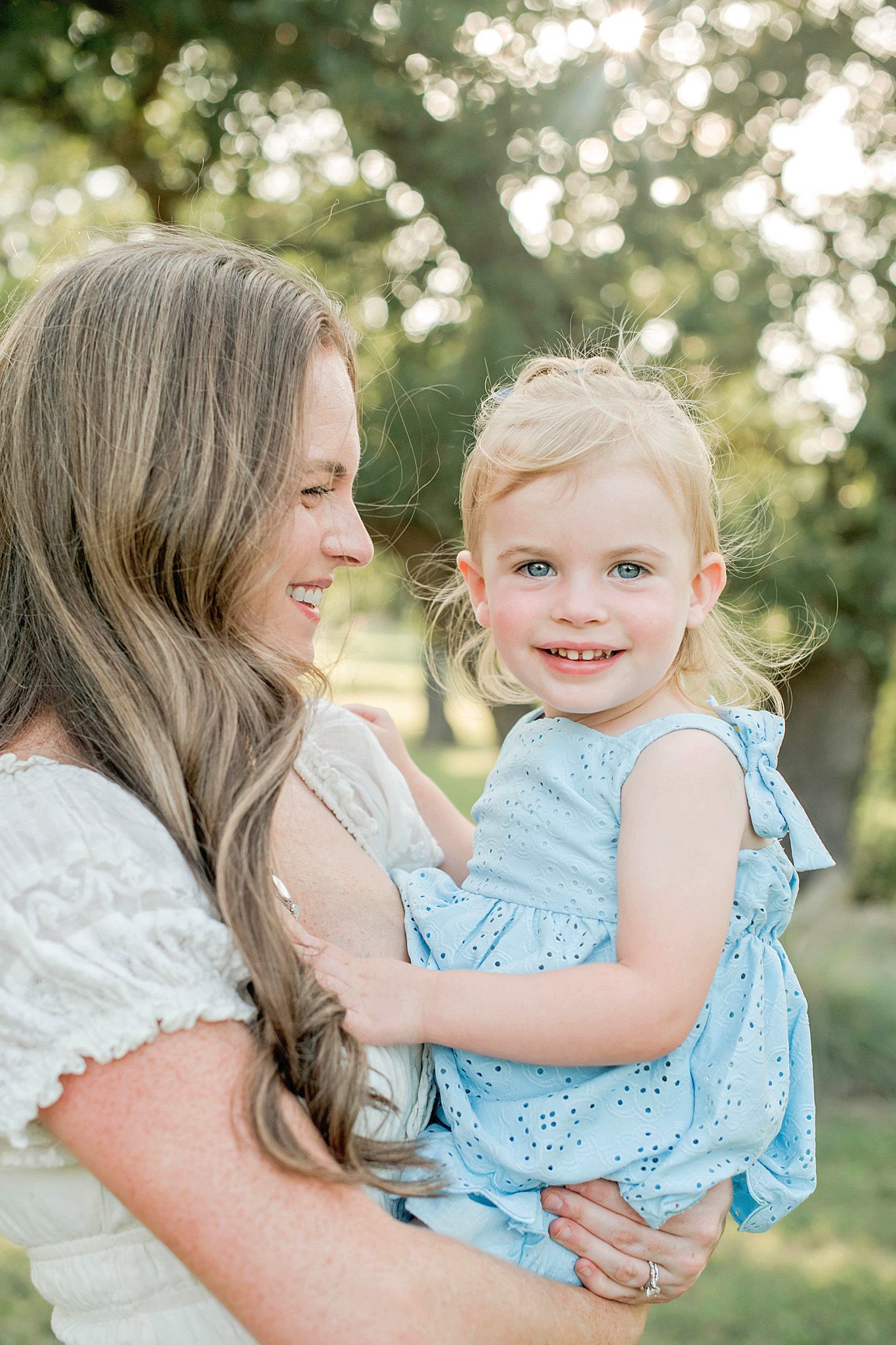 A smiling mother in a white dress holds her happy toddler girl in a blue dress outdoors in golden sunlight.