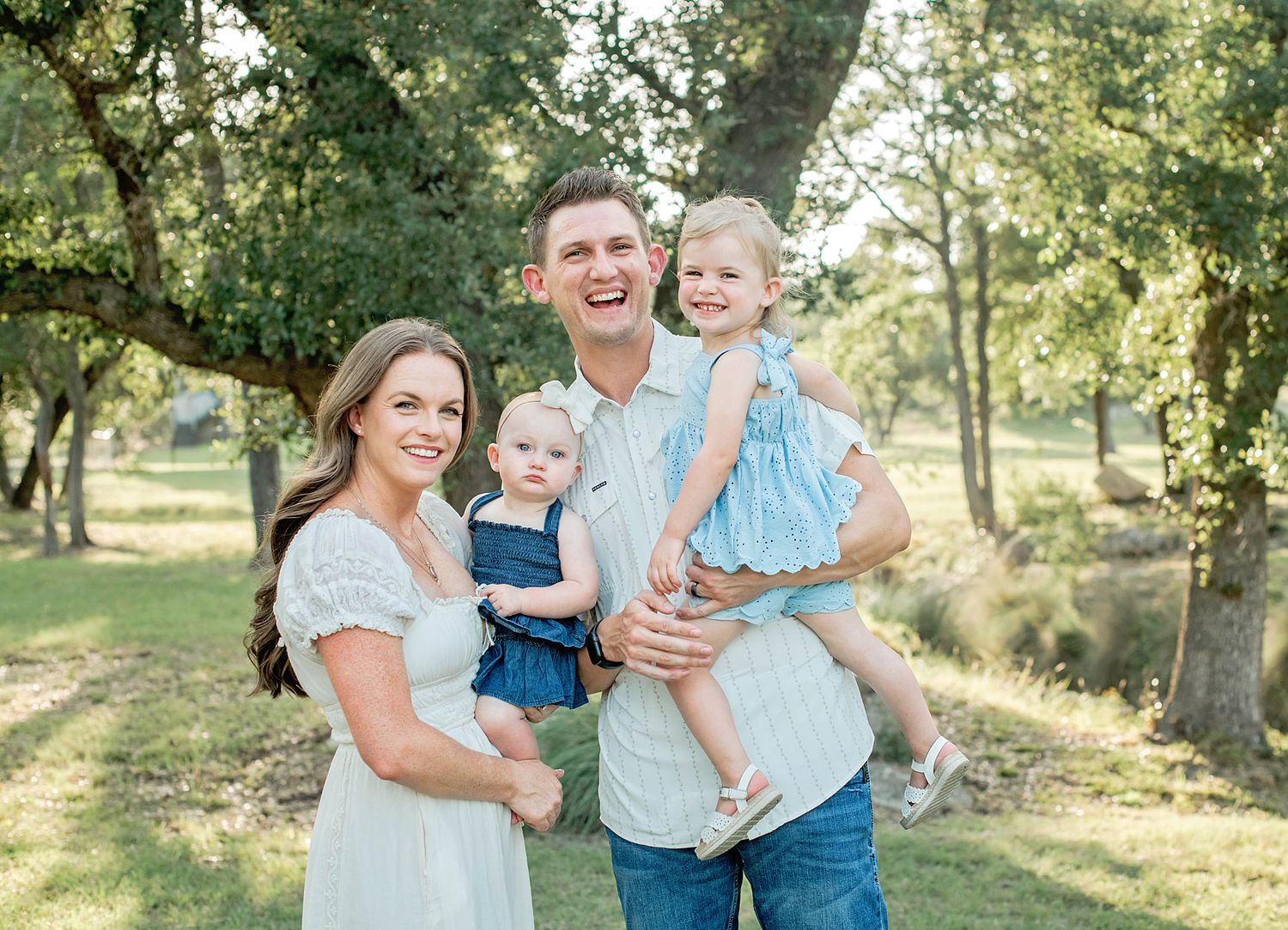 Happy family of four posing outdoors among green trees, parents holding two young daughters in matching blue outfits.