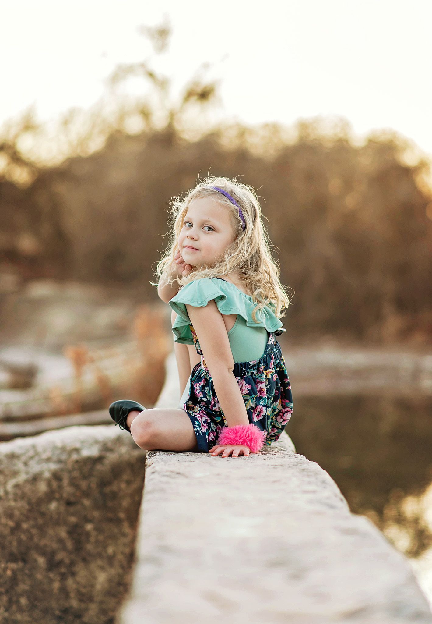A child in a floral dress and mint cardigan plays on large rocks at sunset near a lake.