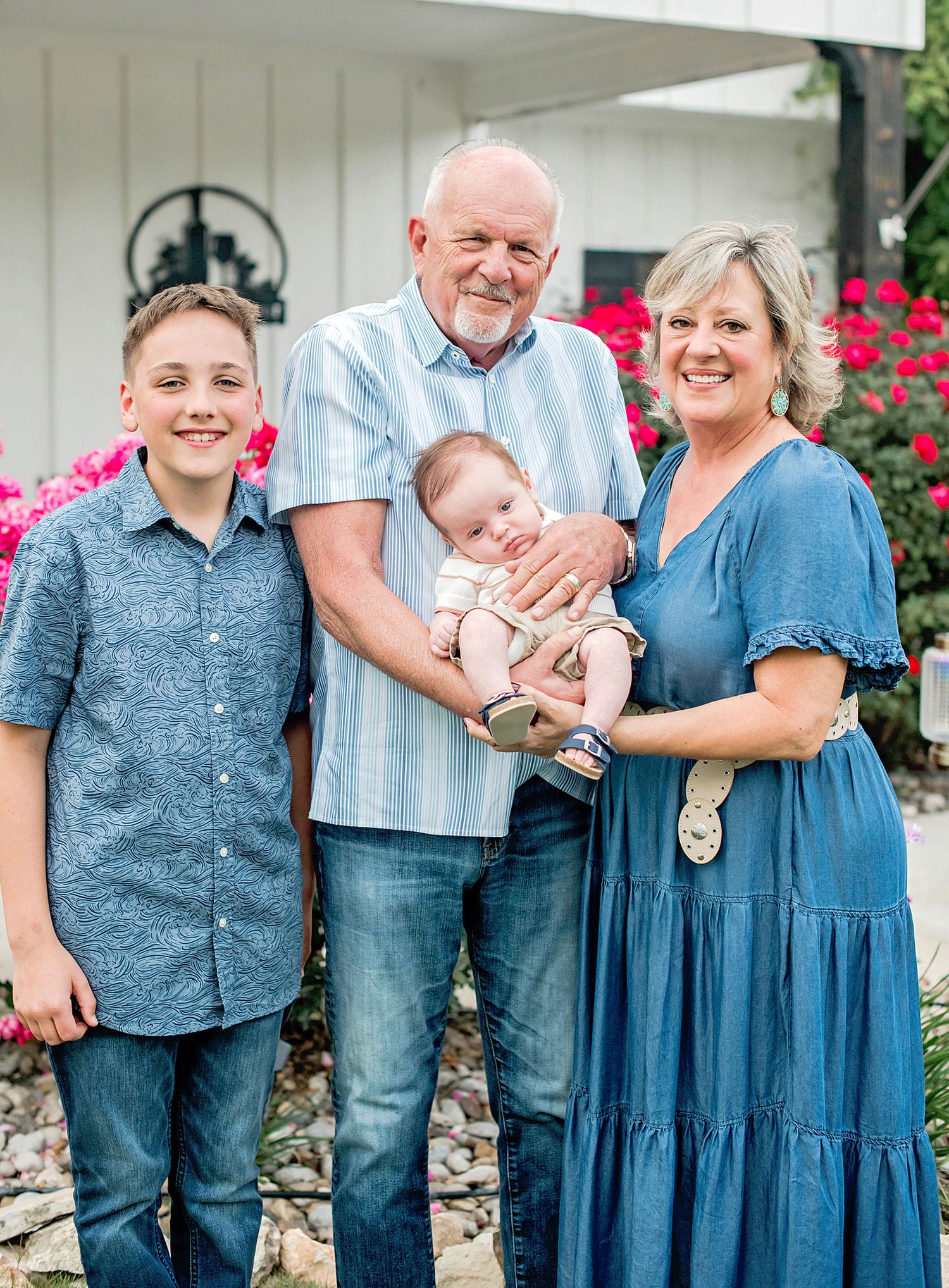 Family of four posing outdoors, grandparents holding a newborn baby with a young boy, surrounded by pink flowers.
