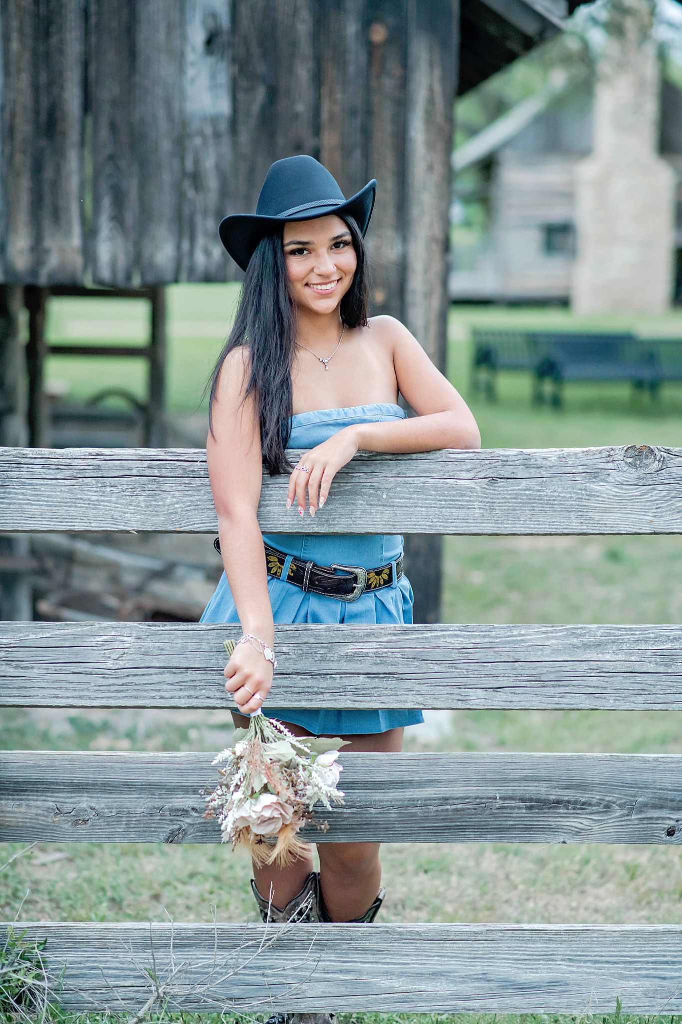 Young woman in blue dress and cowboy hat leans on rustic wooden fence holding wildflowers at a farm.
