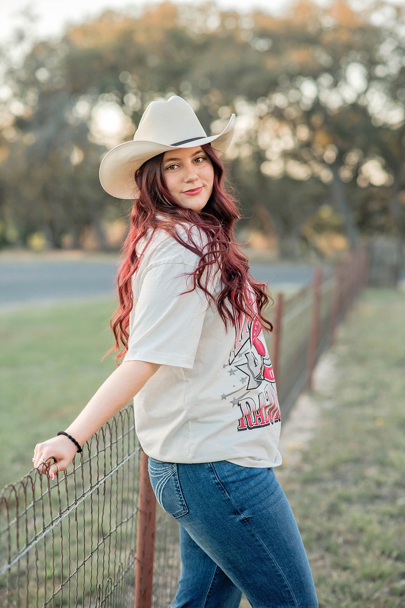 Young woman with red hair wearing cowboy hat and jeans leaning on fence outdoors.