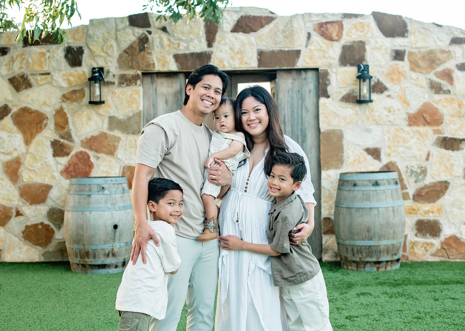 Happy family of five posing outdoors near stone wall and wooden barrels, wearing coordinated neutral outfits.