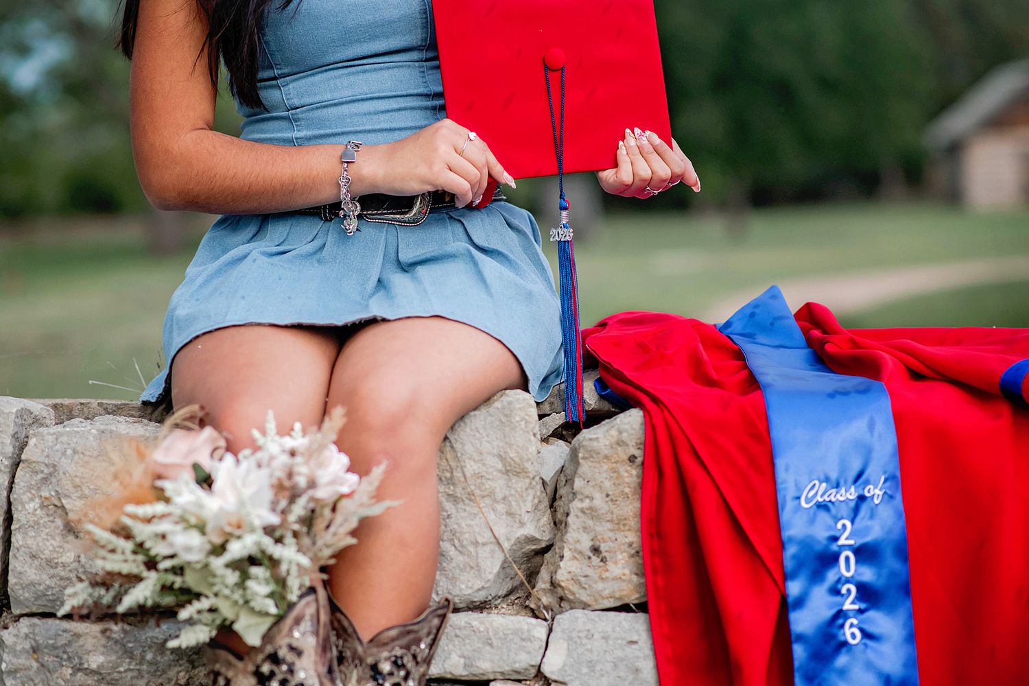 Graduate sits on stone wall holding red cap with Class of 2025 sash and gown, wearing blue dress and boots.