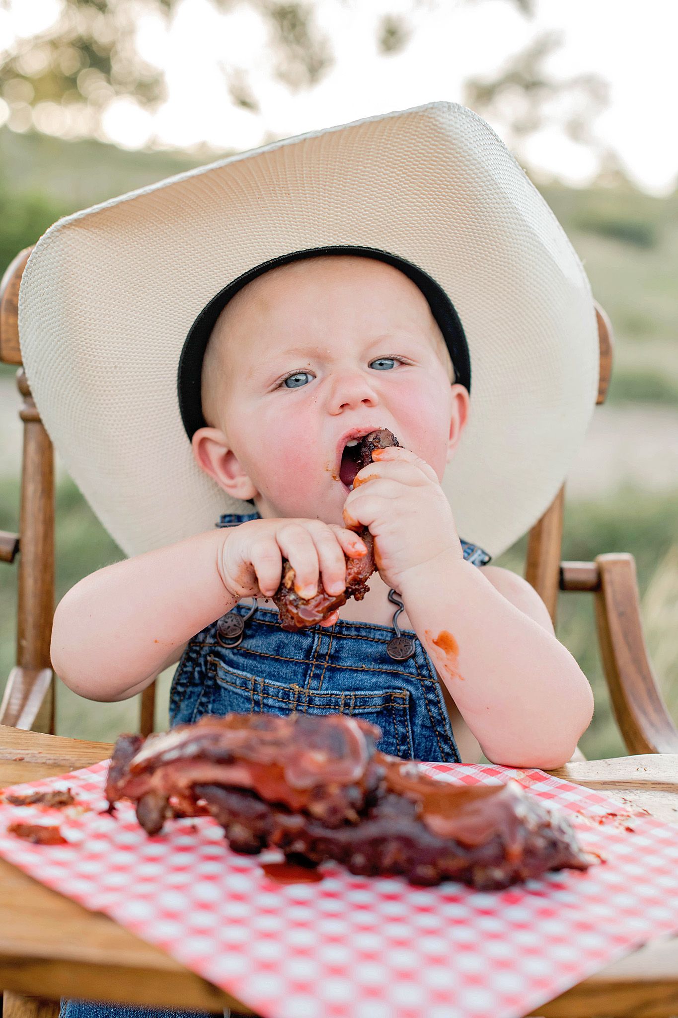 Adorable baby in cowboy hat and denim overalls eating BBQ ribs at outdoor table with red checkered cloth.