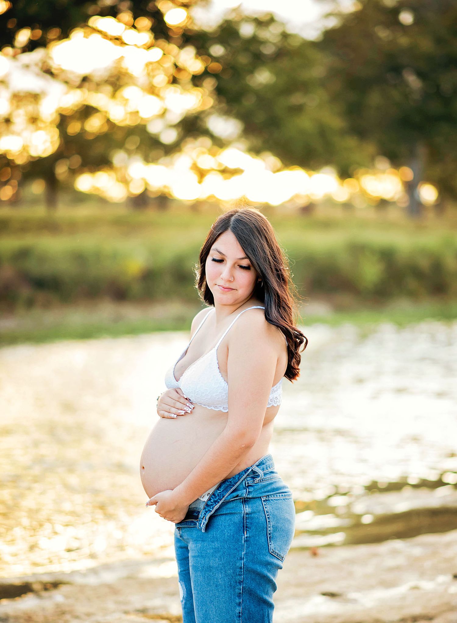 Maternity photoshoot at sunset with woman in denim overalls standing by water's edge in golden light.