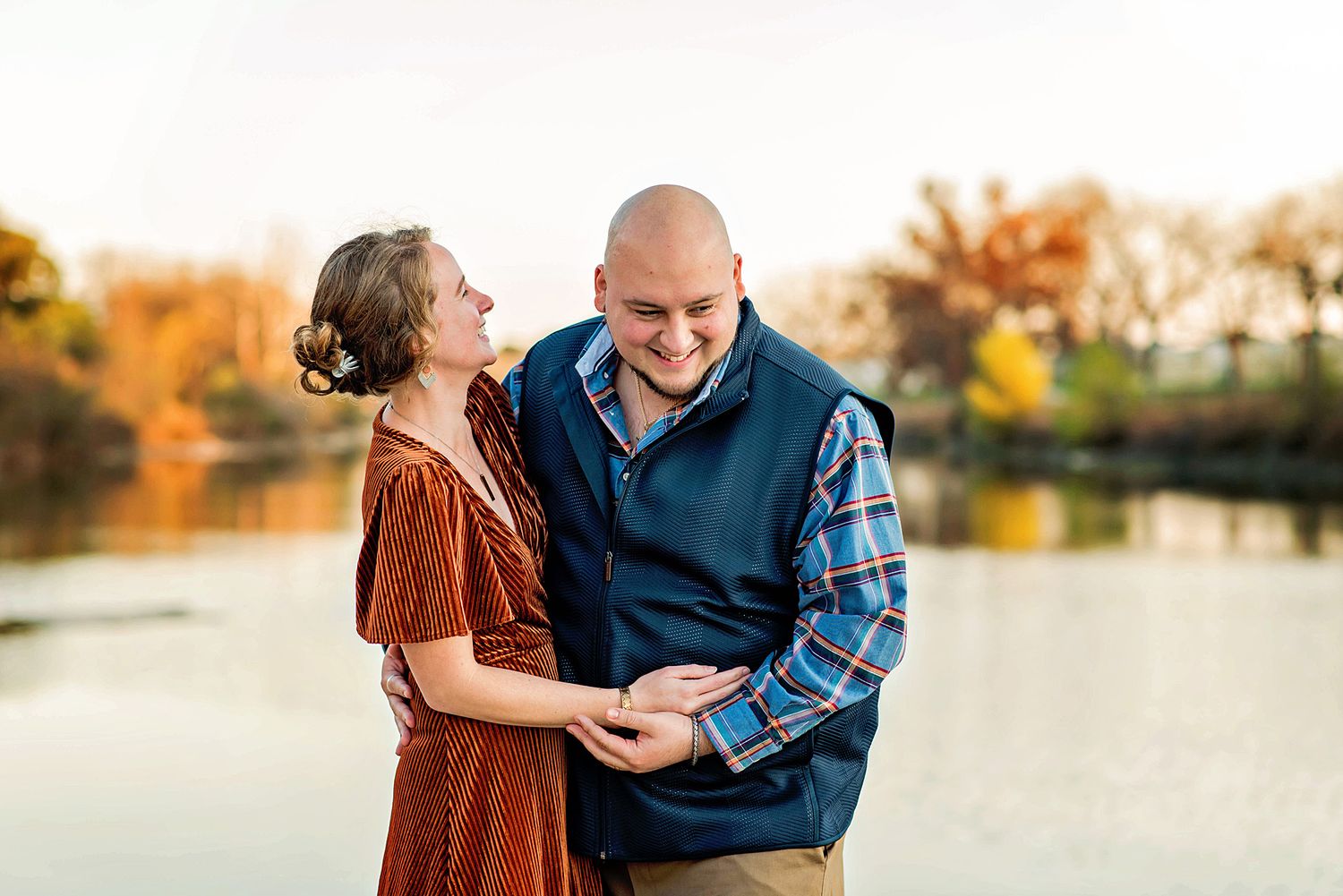 A couple laughs together while embracing by a lake during autumn with colorful fall foliage in the background.