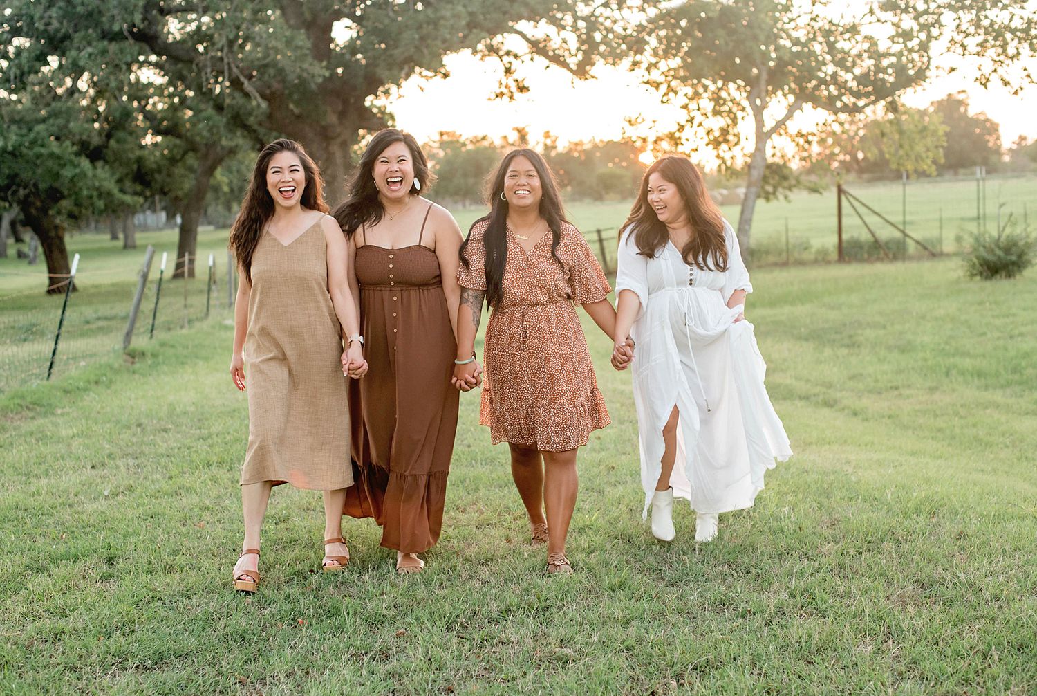 Four women holding hands walk through a sunlit green field at golden hour wearing earth-toned and white dresses.