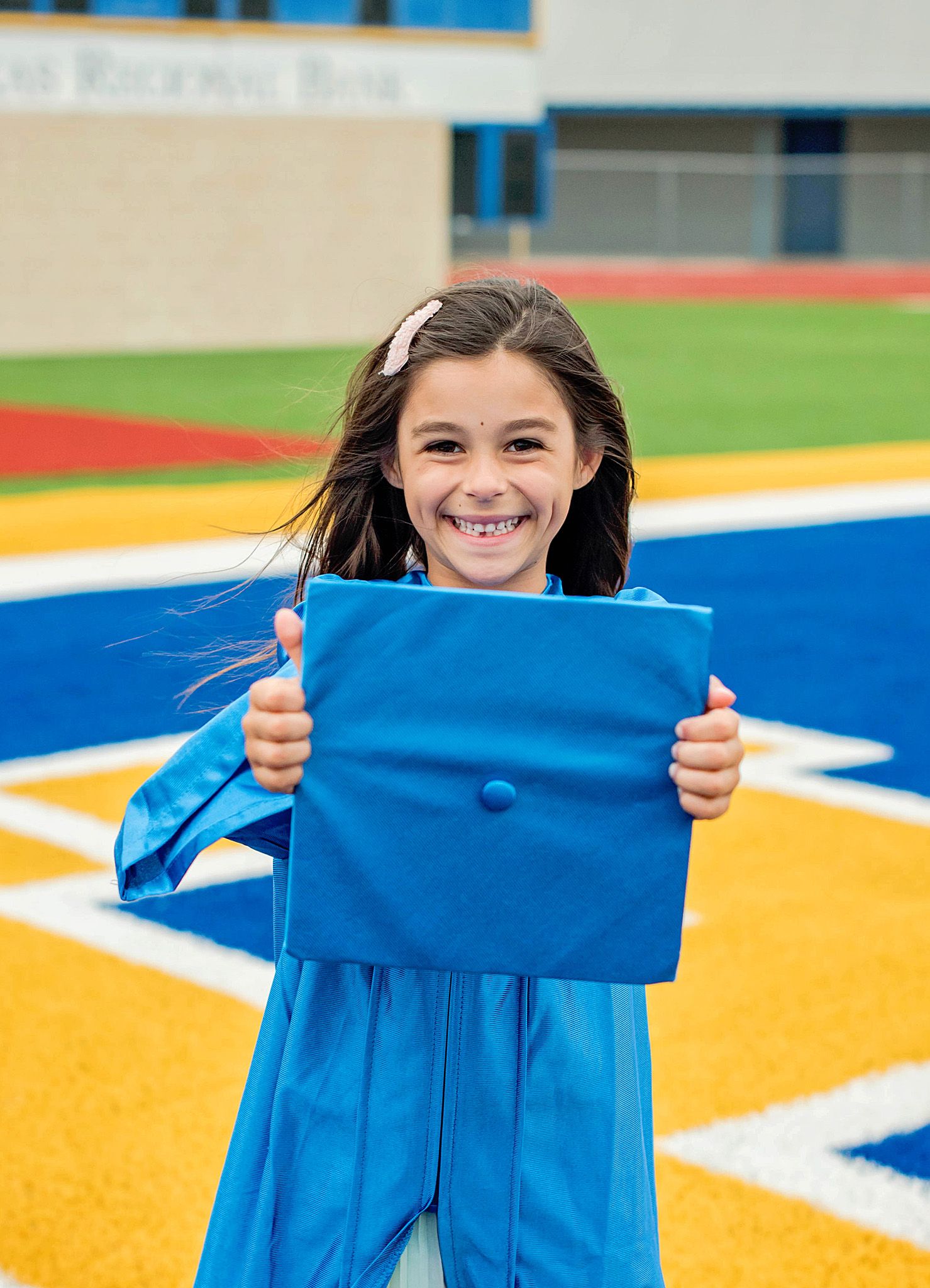 Young graduate in bright blue gown holds up diploma cover on colorful sports field.