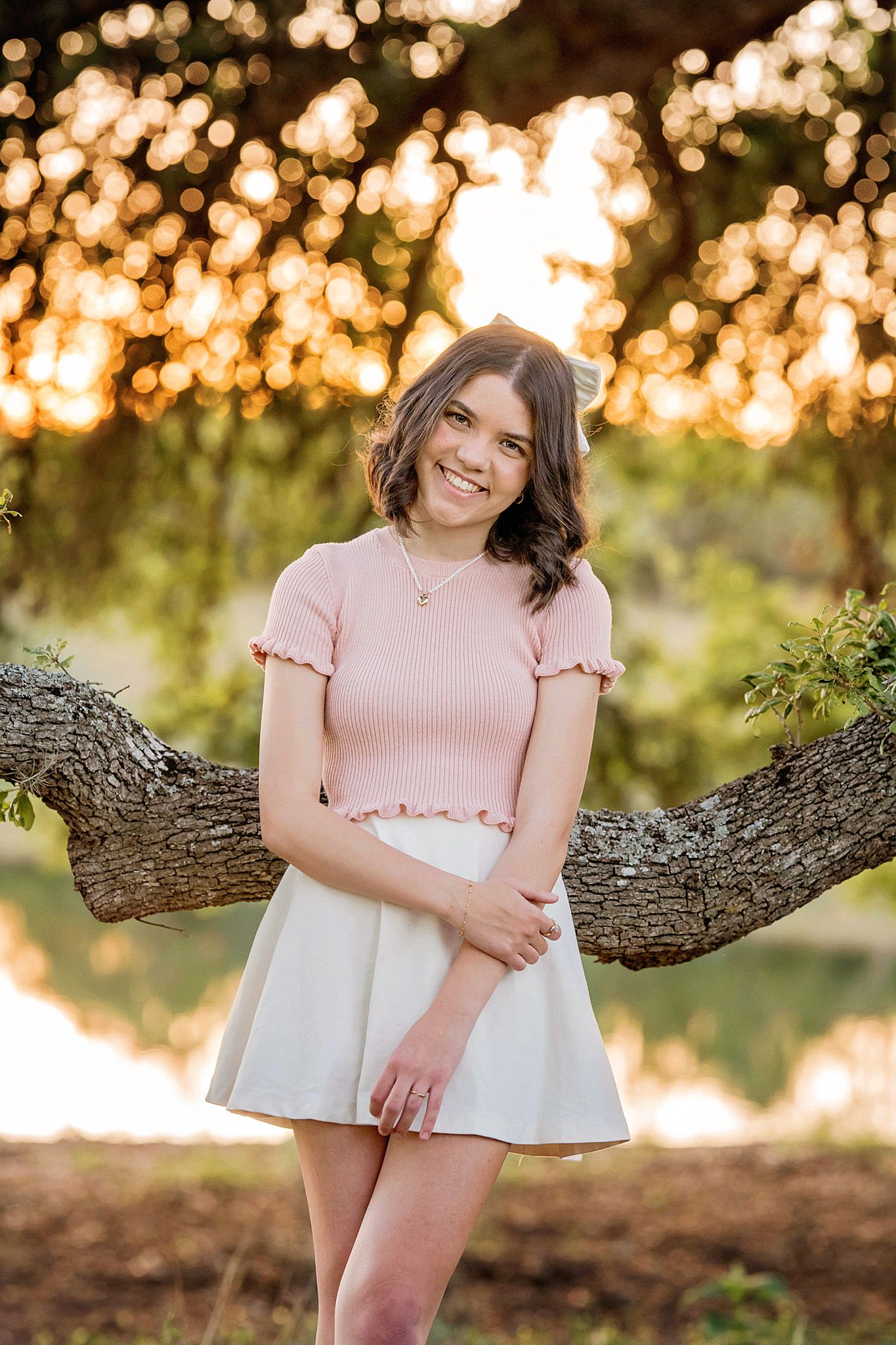 Young woman in pink crop top and white skirt smiling outdoors during golden hour near a tree.