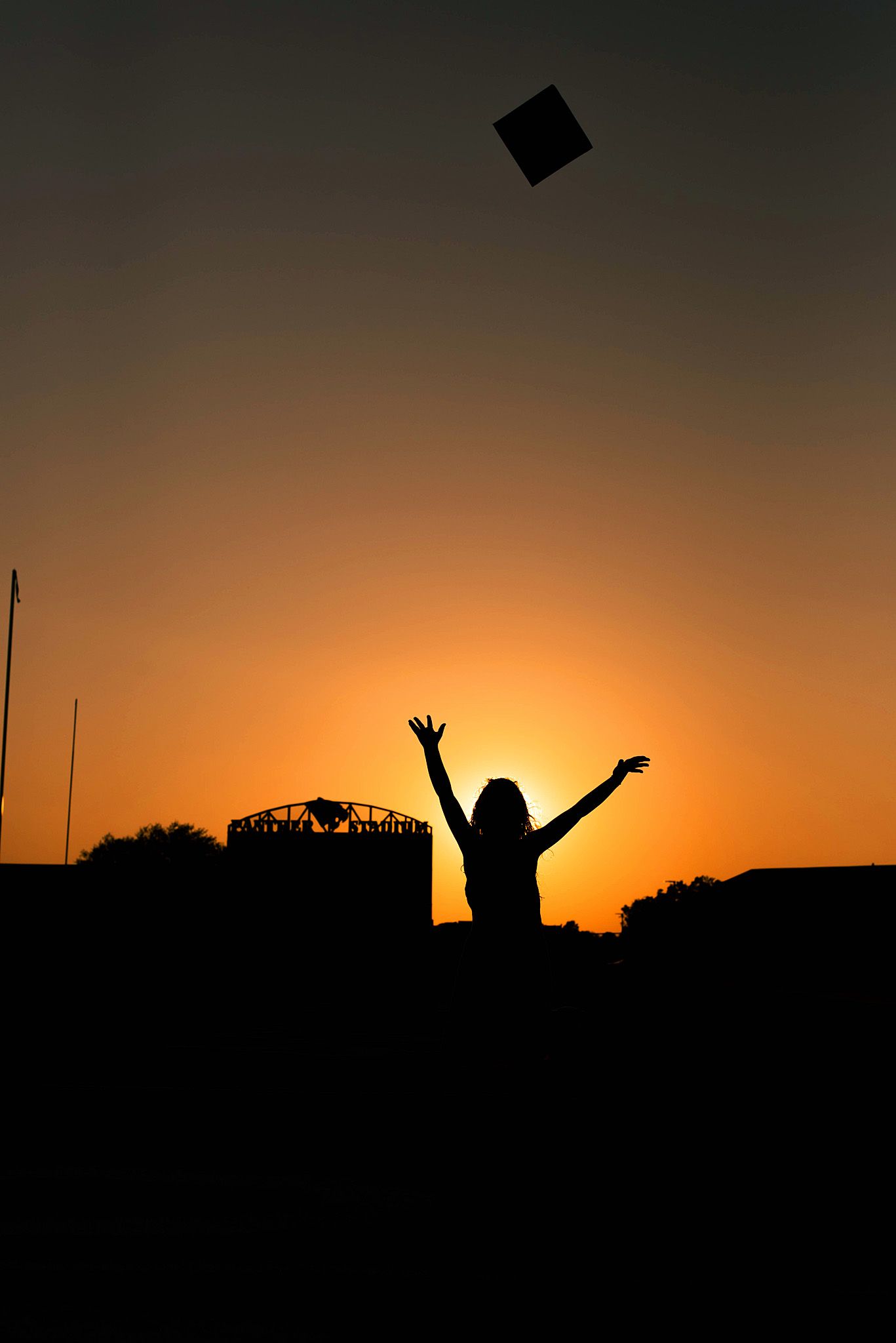 Silhouette of a graduate tossing cap at sunset with arms raised in celebration against an orange sky.