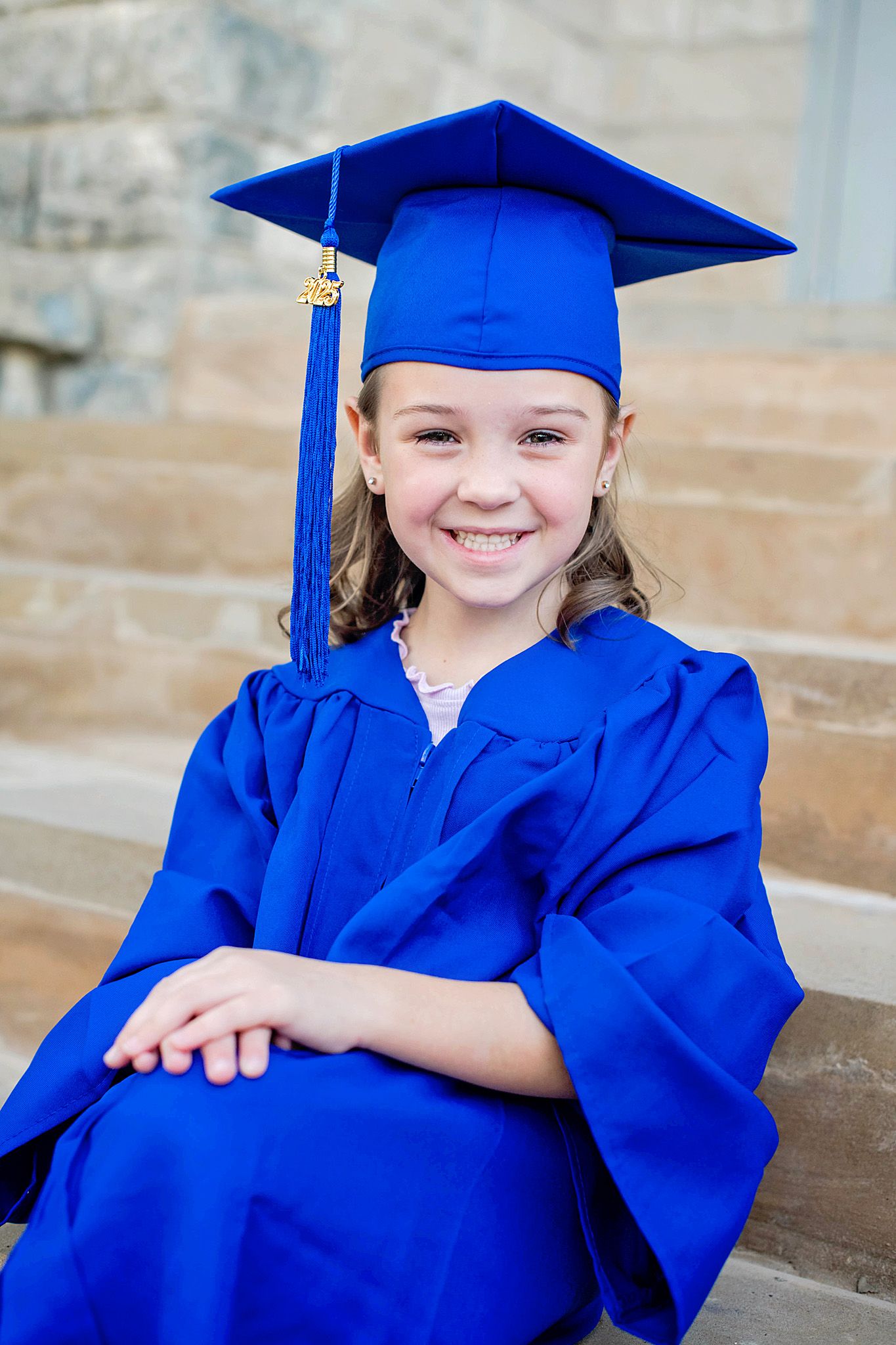 Young girl smiling in blue graduation cap and gown, sitting on steps during kindergarten graduation ceremony.