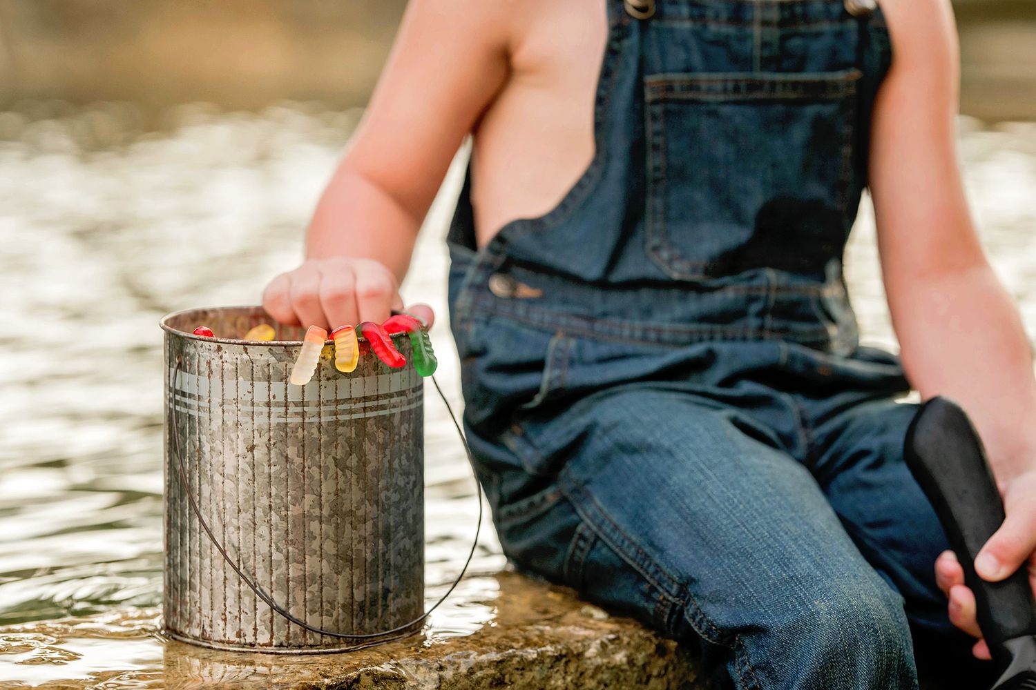 Young child in denim overalls sits by water holding a fishing rod next to a metal bait bucket.