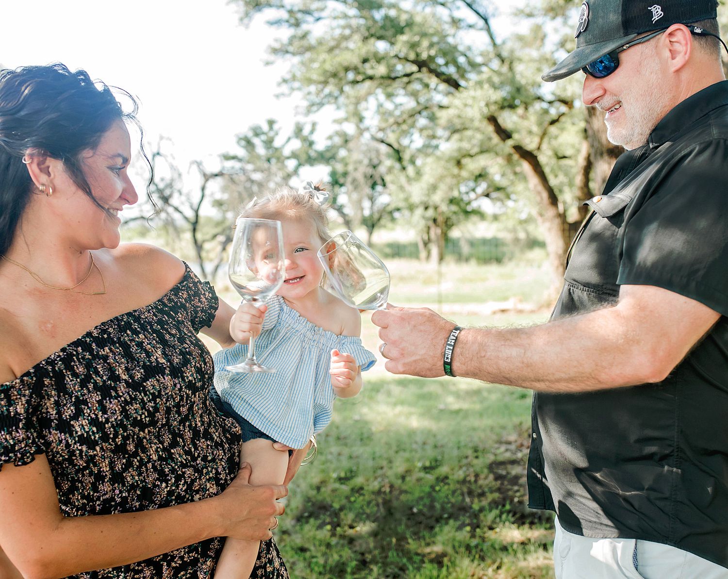 Family toasting with wine glasses outdoors, smiling couple holding young child among green trees on sunny day.