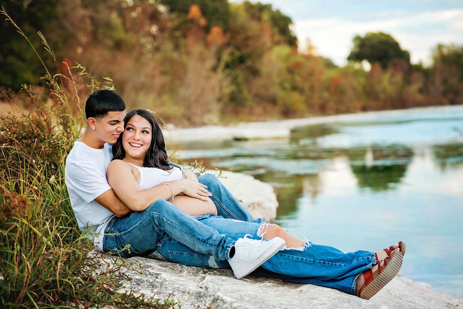 Couple relaxing together on rocky shoreline by tranquil lake during sunset photo session.