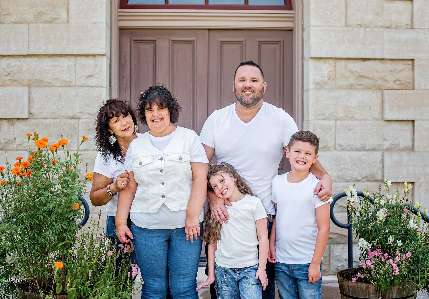 A family stands together smiling in front of a stone building with orange and pink flowers blooming in the garden in Blanco, TX.