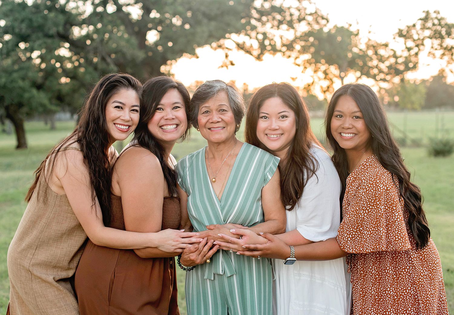 Five women smiling together outdoors at golden hour, posing in a sunny park setting for a family portrait.