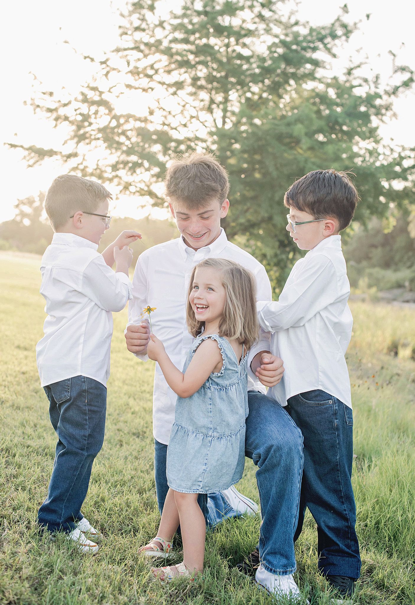 Four children laughing and playing together in a sunny green field, wearing white shirts and jeans.