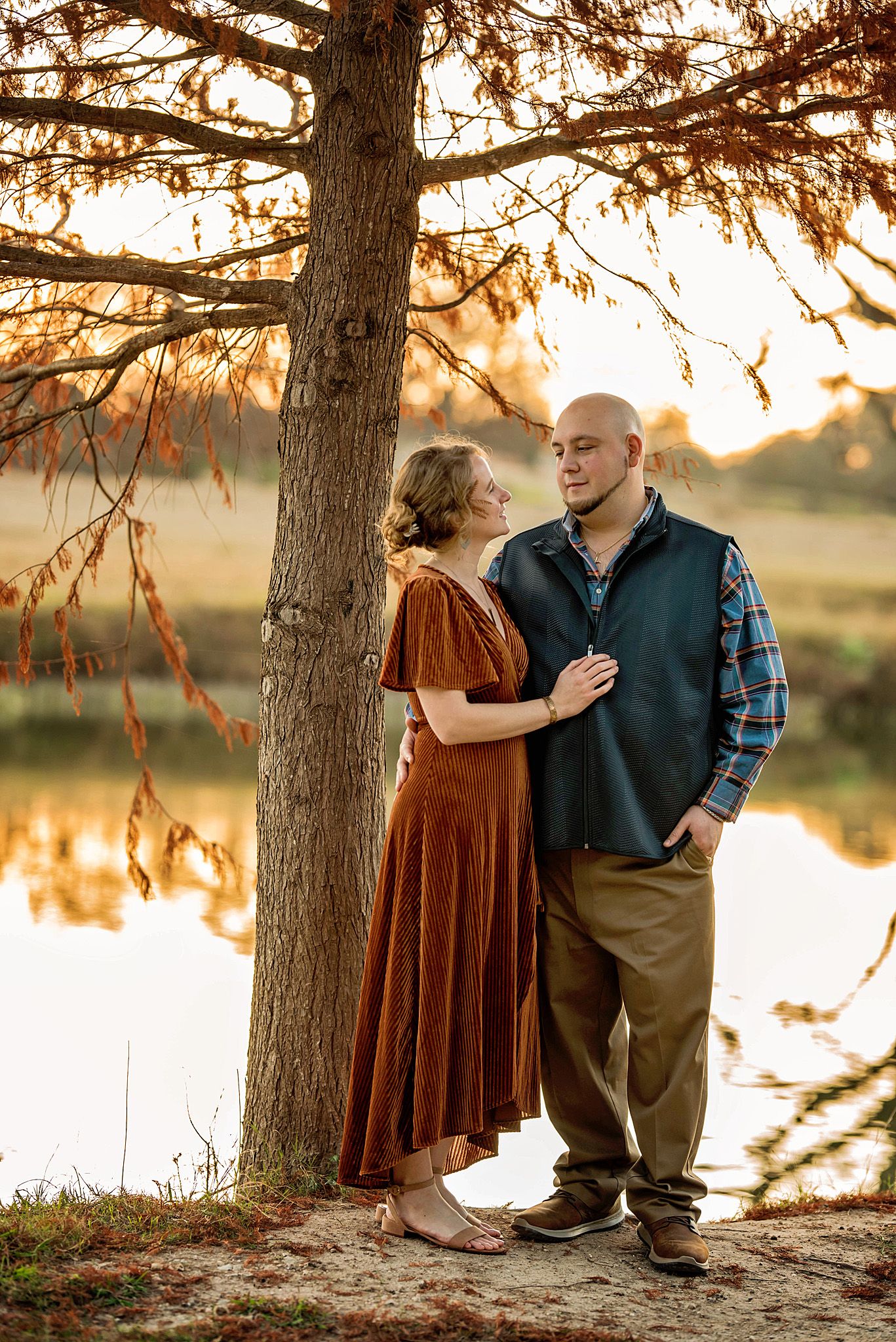 Couple embraces near a lake at sunset with golden light filtering through tree branches.