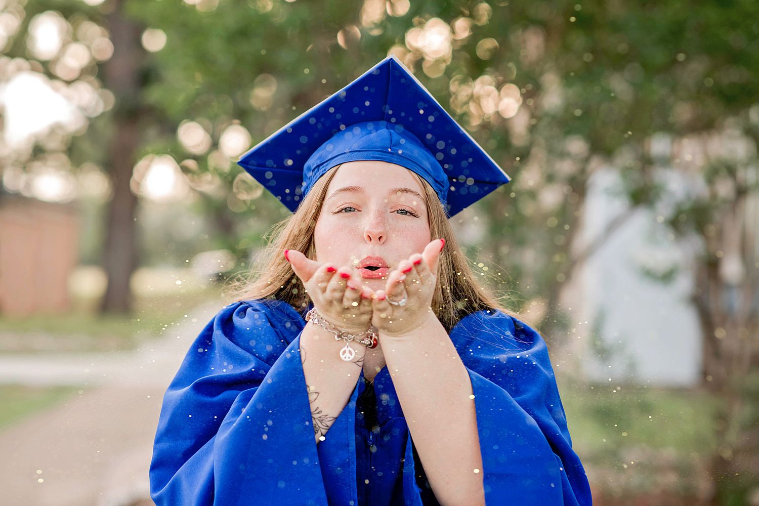 A young woman in a blue graduation cap and gown blows glitter from her hands outdoors.