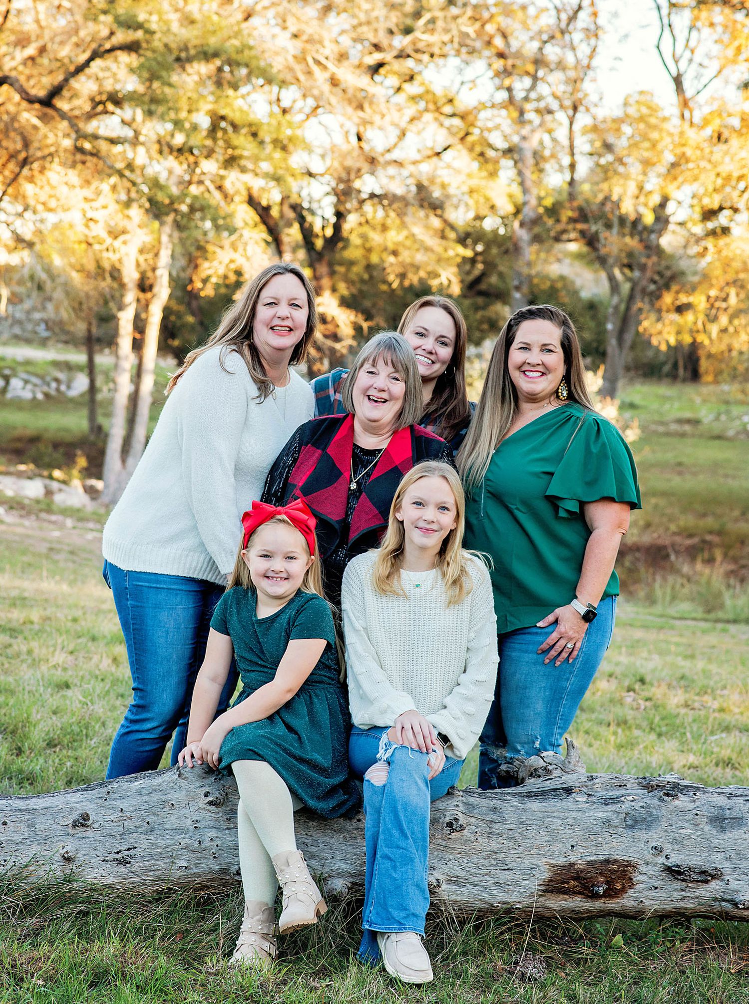A joyful multigenerational family of women and girls posing outdoors on a fallen log surrounded by autumn trees.