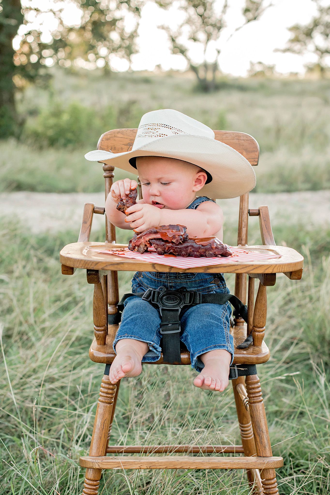 Baby sits in a wooden high chair eating bbq ribs on a grassy outdoor field in blue overalls and bare feet.