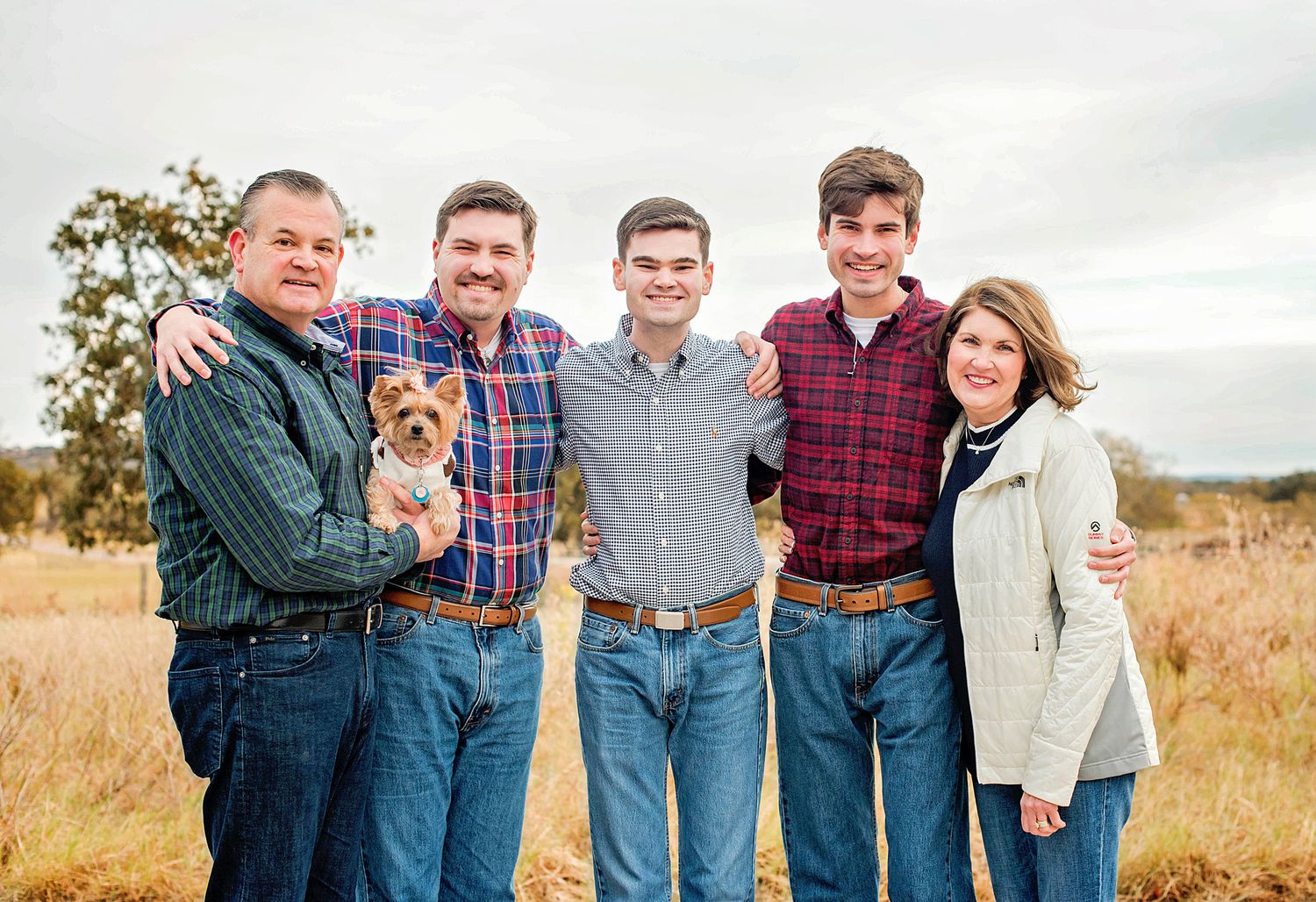 Family of five posing outdoors in a field, smiling together while holding a small dog on a sunny day.