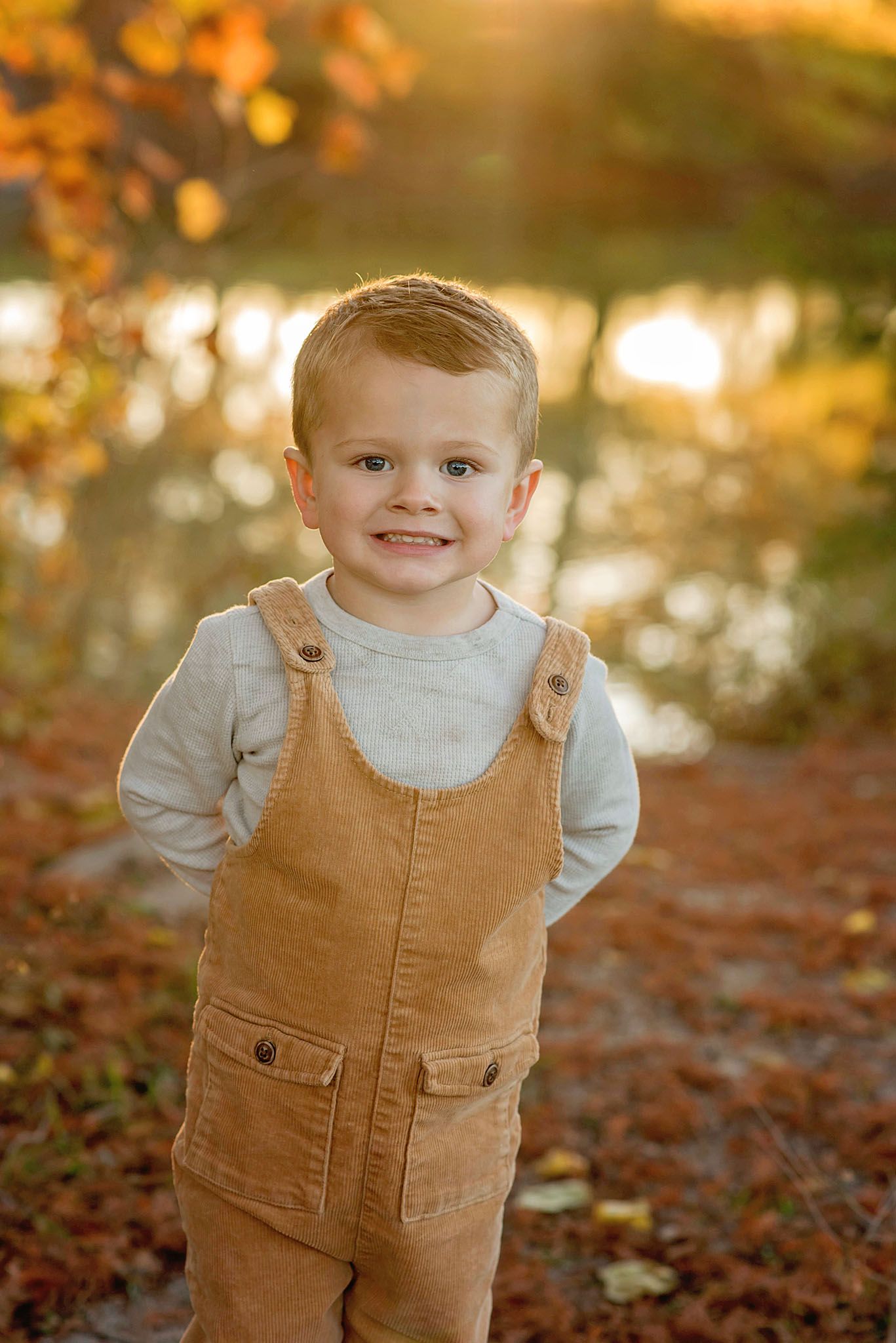 A toddler in tan corduroy overalls and white shirt smiles during an autumn photoshoot with golden leaves in background.