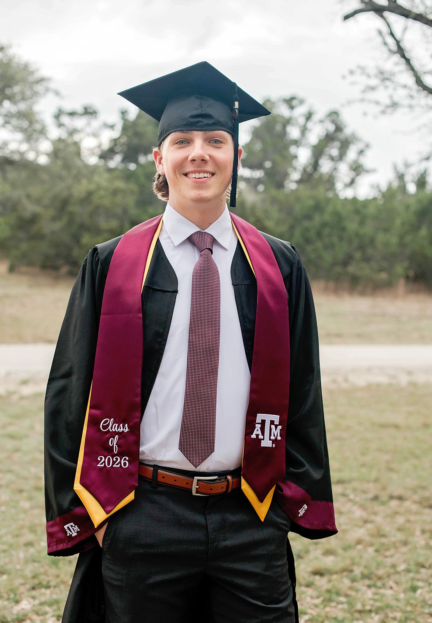 Texas A&M student smiling in graduation cap, gown, and maroon stole reading Class of 2026 outdoors.