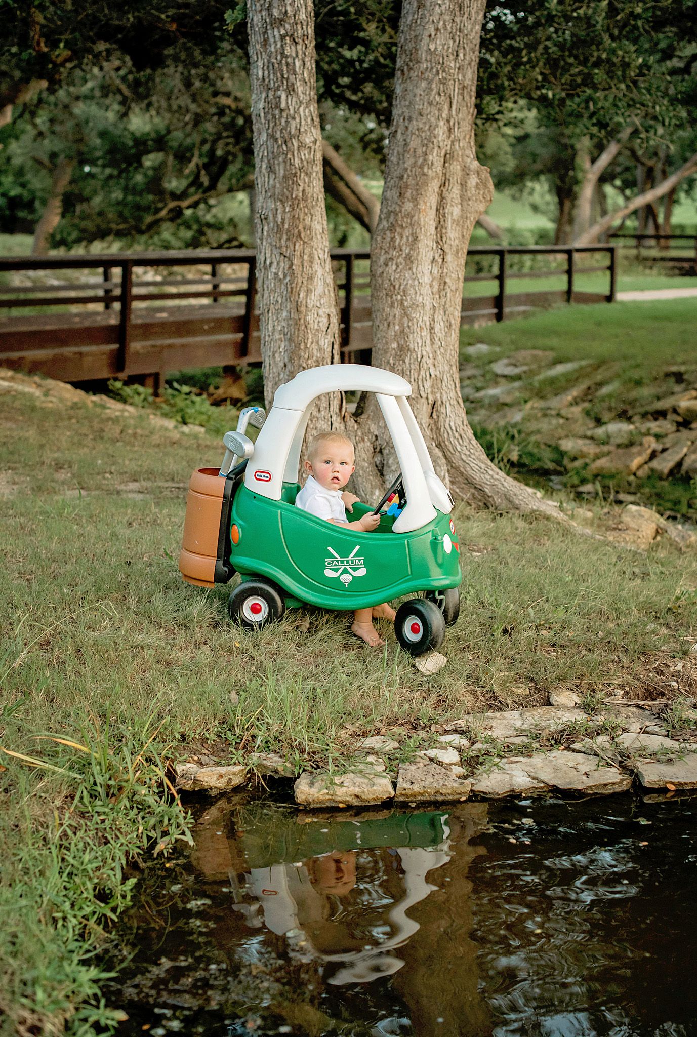 A green and white toy car parked near a pond, surrounded by trees and wooden fencing in a peaceful outdoor setting.