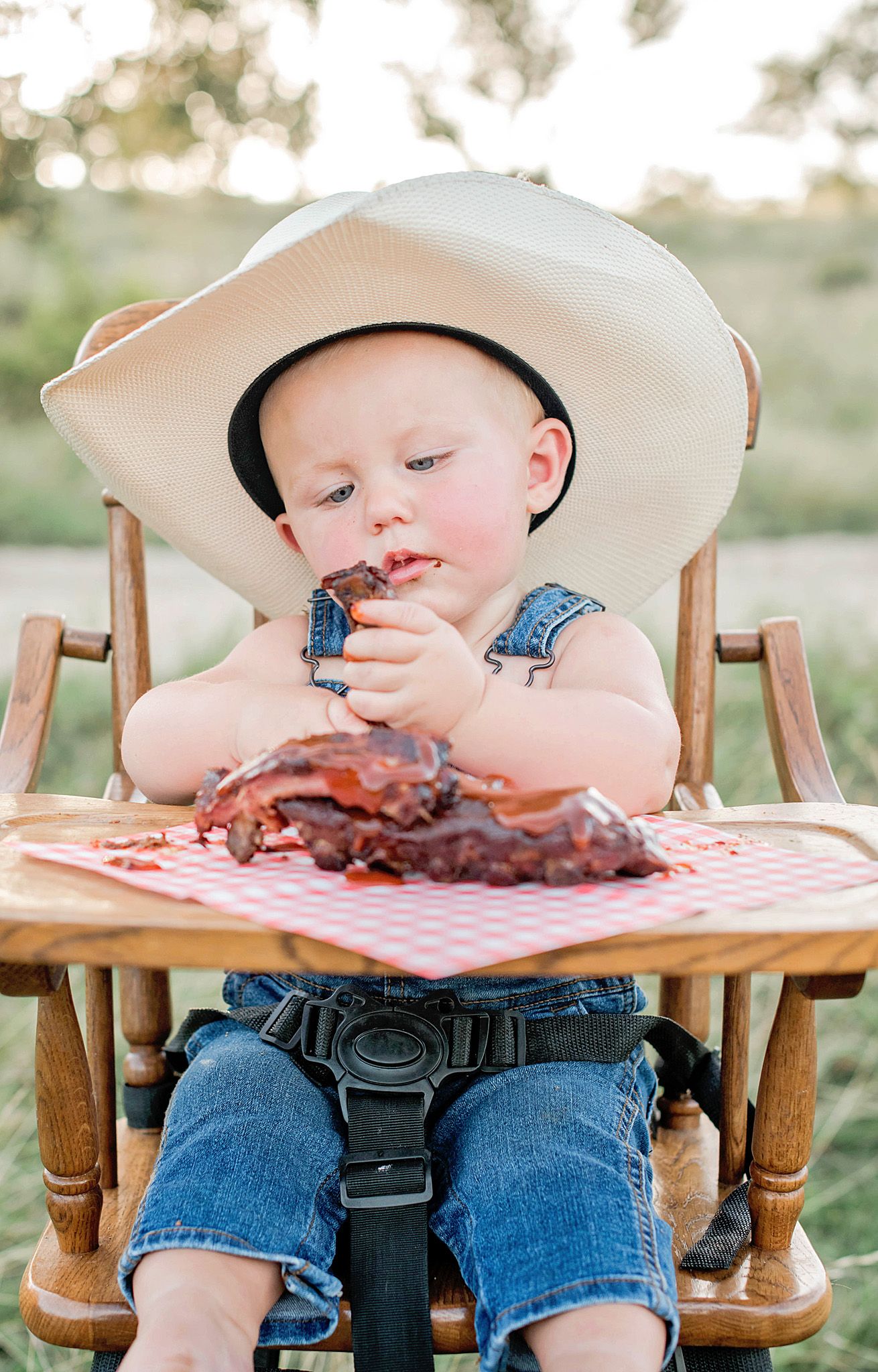 Baby in white cowboy hat and denim overalls sits in wooden highchair eating barbecue ribs at picnic table.
