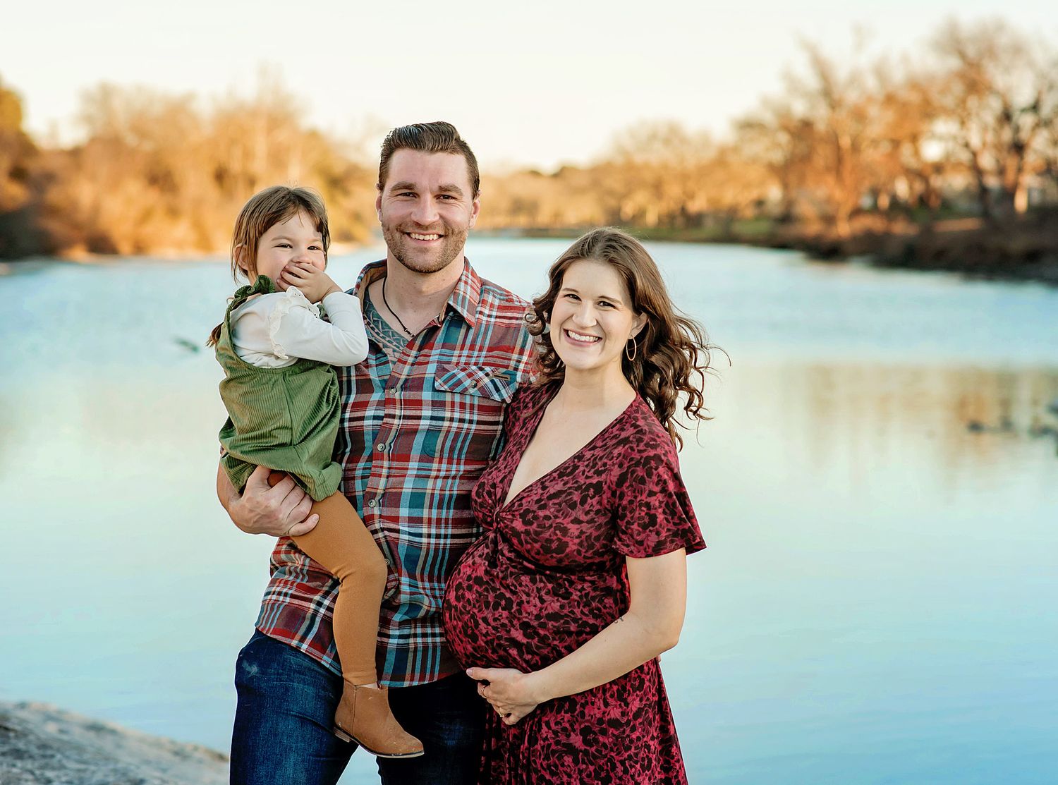 A family in coordinating red and plaid outfits poses together by a scenic autumn lake at sunset.