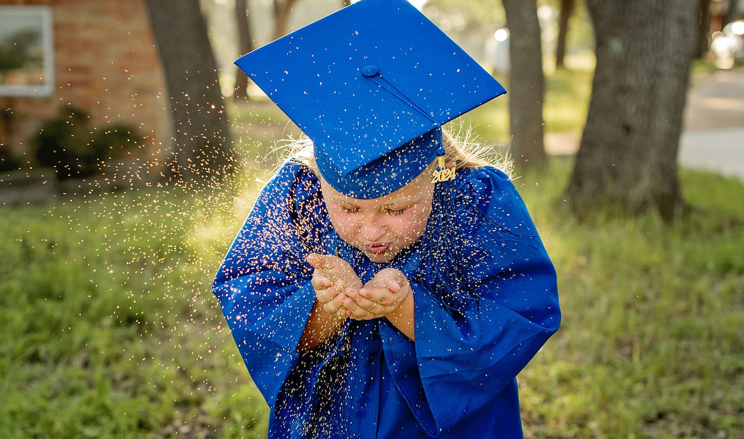 Graduate in blue cap and gown throws glitter in celebration during outdoor graduation photoshoot.