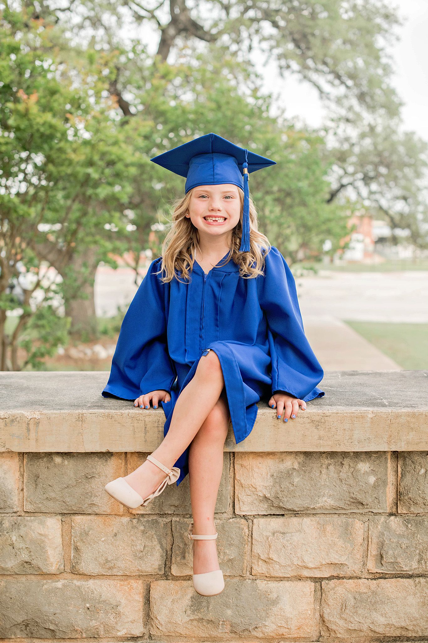 A young girl smiles in a blue graduation cap and gown, sitting on a stone wall outdoors.