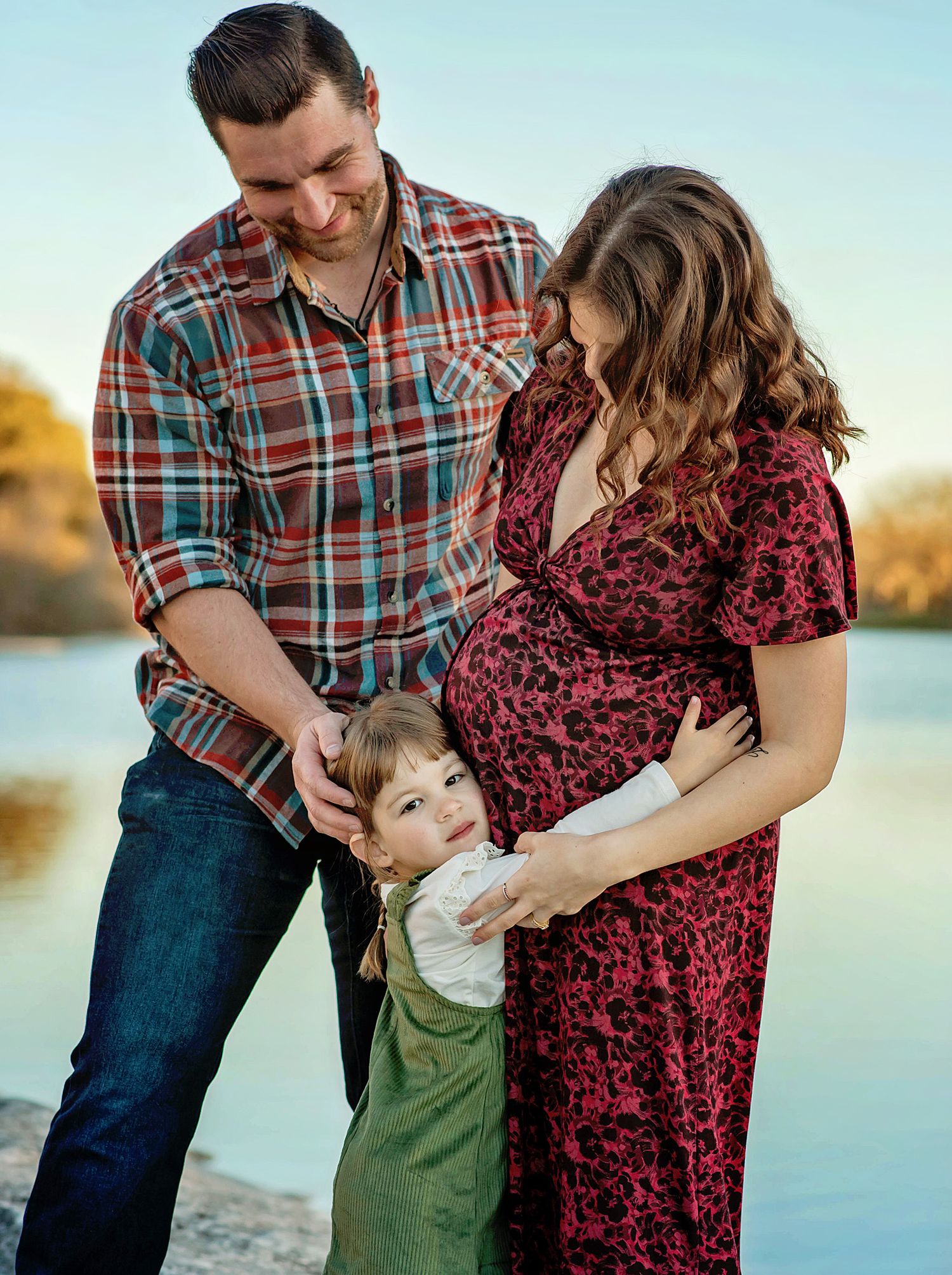 Family shares tender moments together by a serene lake during sunset, wearing plaid and burgundy outfits.