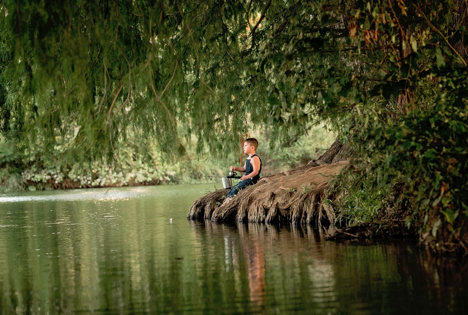 Boy sitting on rocks by a calm river surrounded by lush green trees, enjoying a peaceful moment in nature.