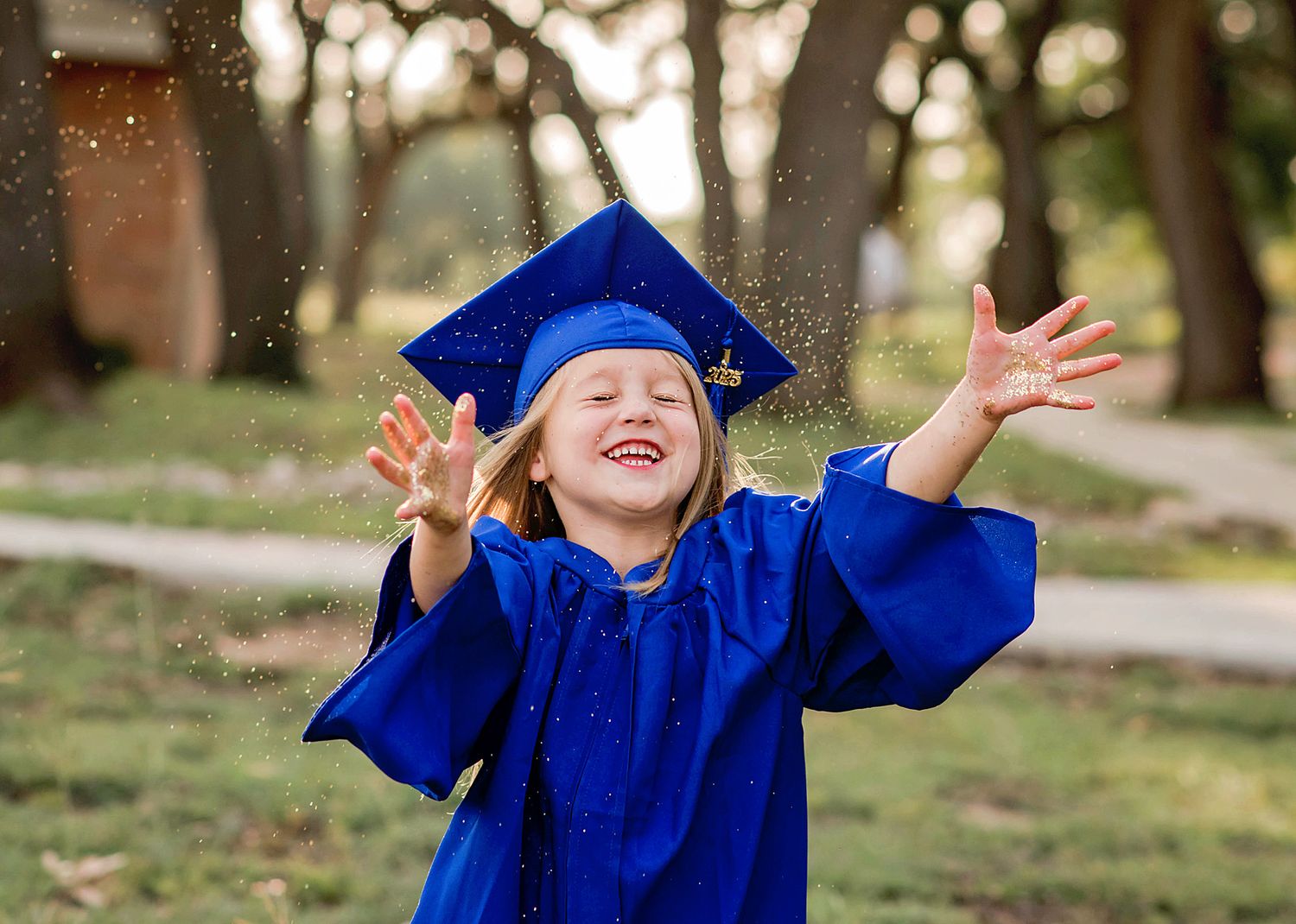A graduate in blue cap and gown celebrates joyfully in an outdoor setting during a graduation ceremony.