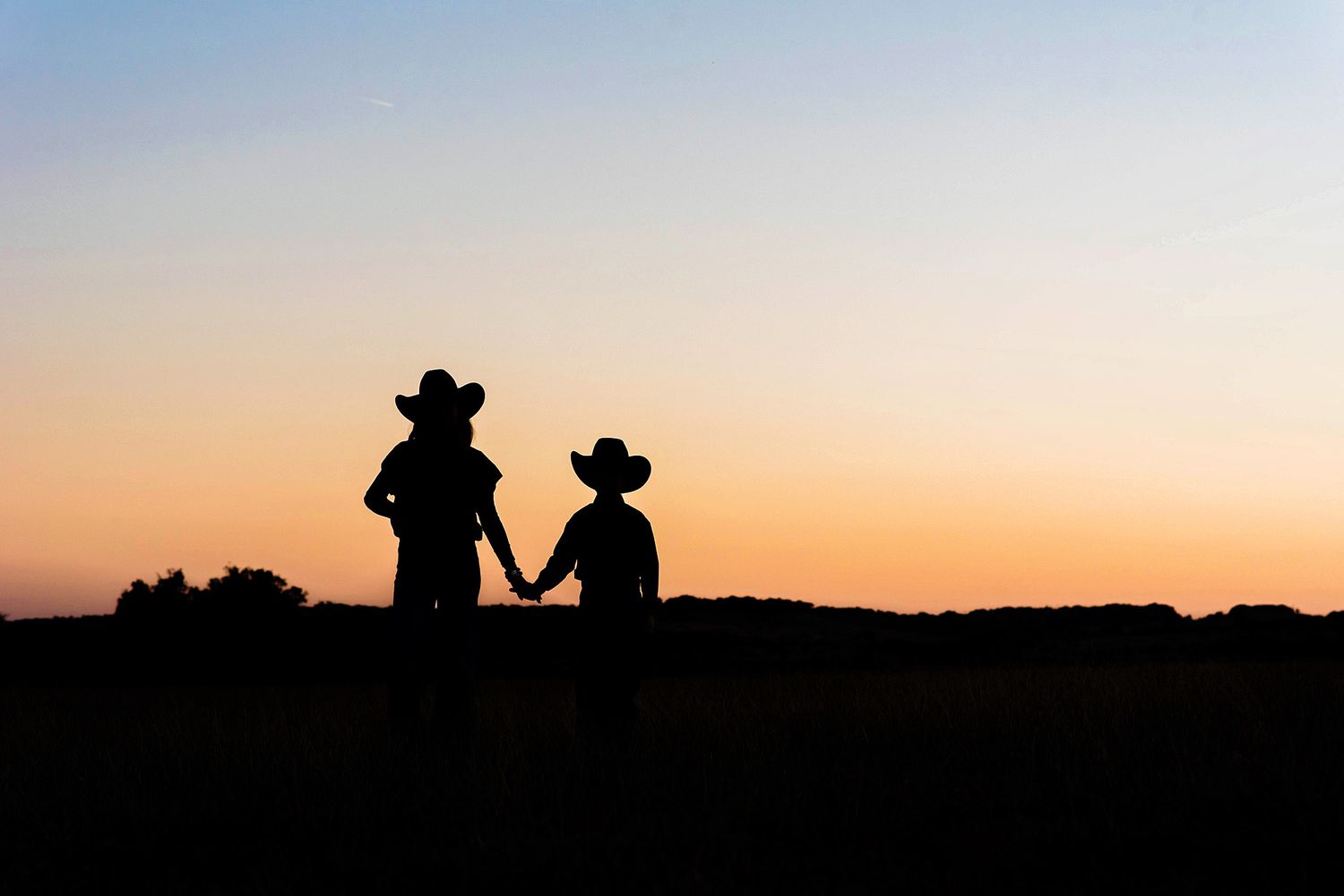 Silhouetted figures and horses stand against a vibrant orange sunset sky in a western landscape in Hye, TX.