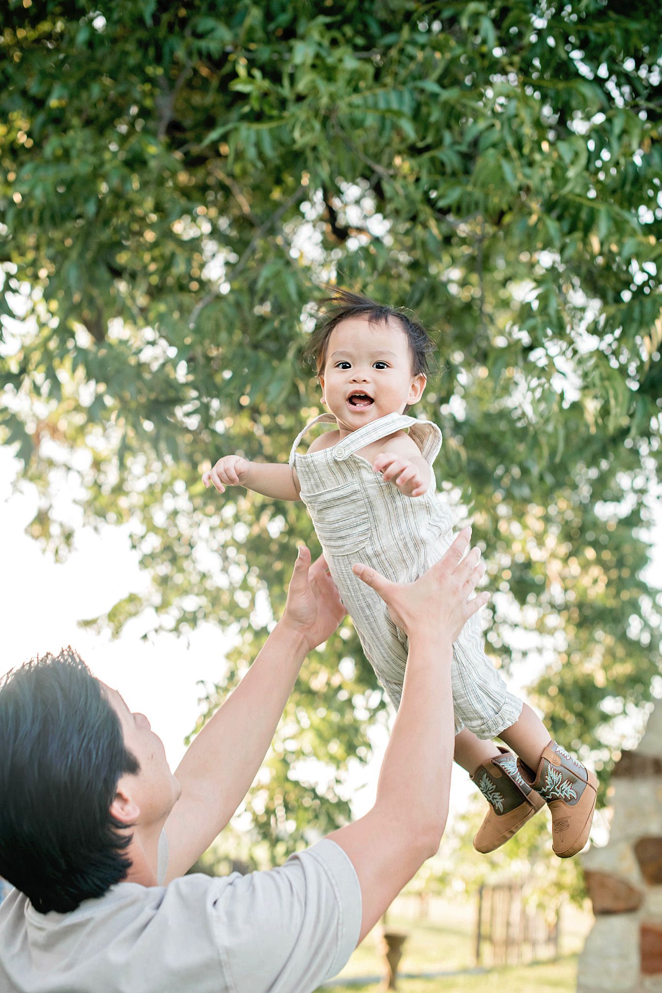 A joyful parent lifts a smiling baby in a white romper outdoors beneath lush green trees.