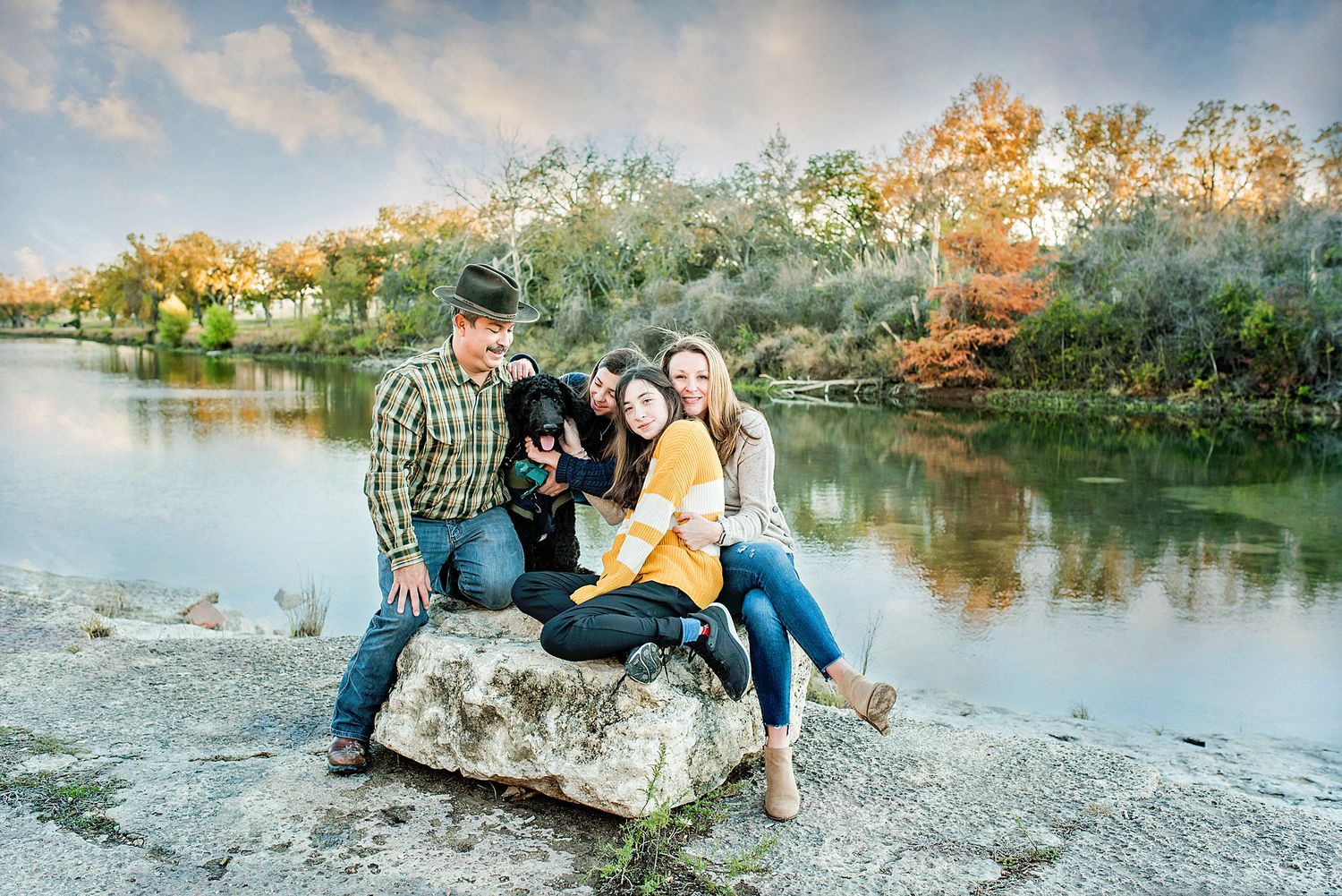 Family of three sitting on a rock by a serene lake with fall foliage, sharing a joyful moment with their black dog.