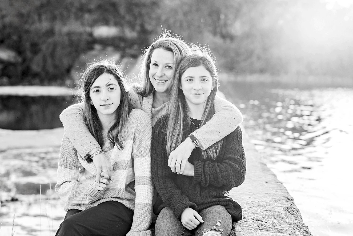 Three friends sitting together on a stone wall near water in black and white photography.