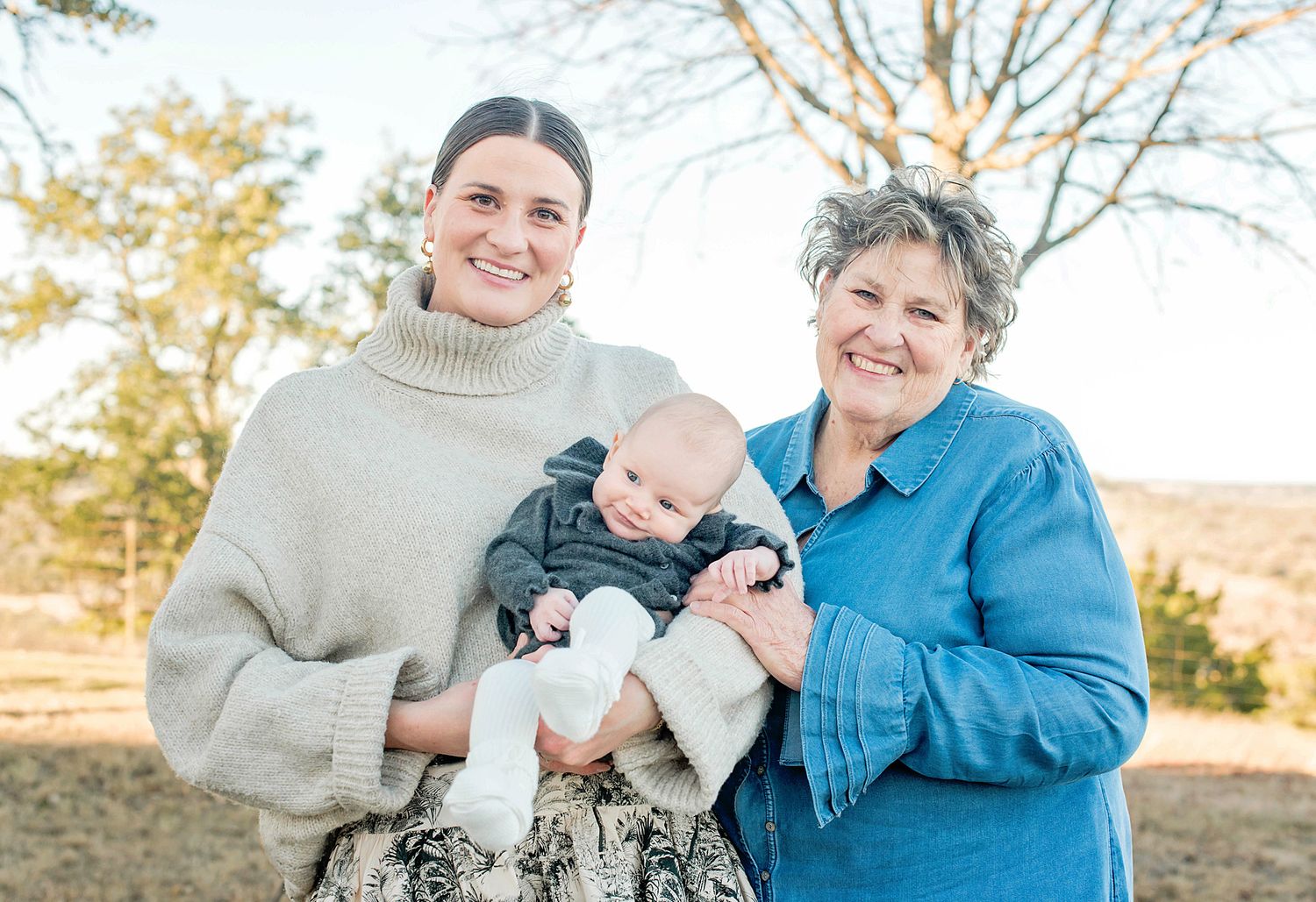 Two women smiling outdoors, one holding a baby in a cozy fall setting with bare trees in the background.