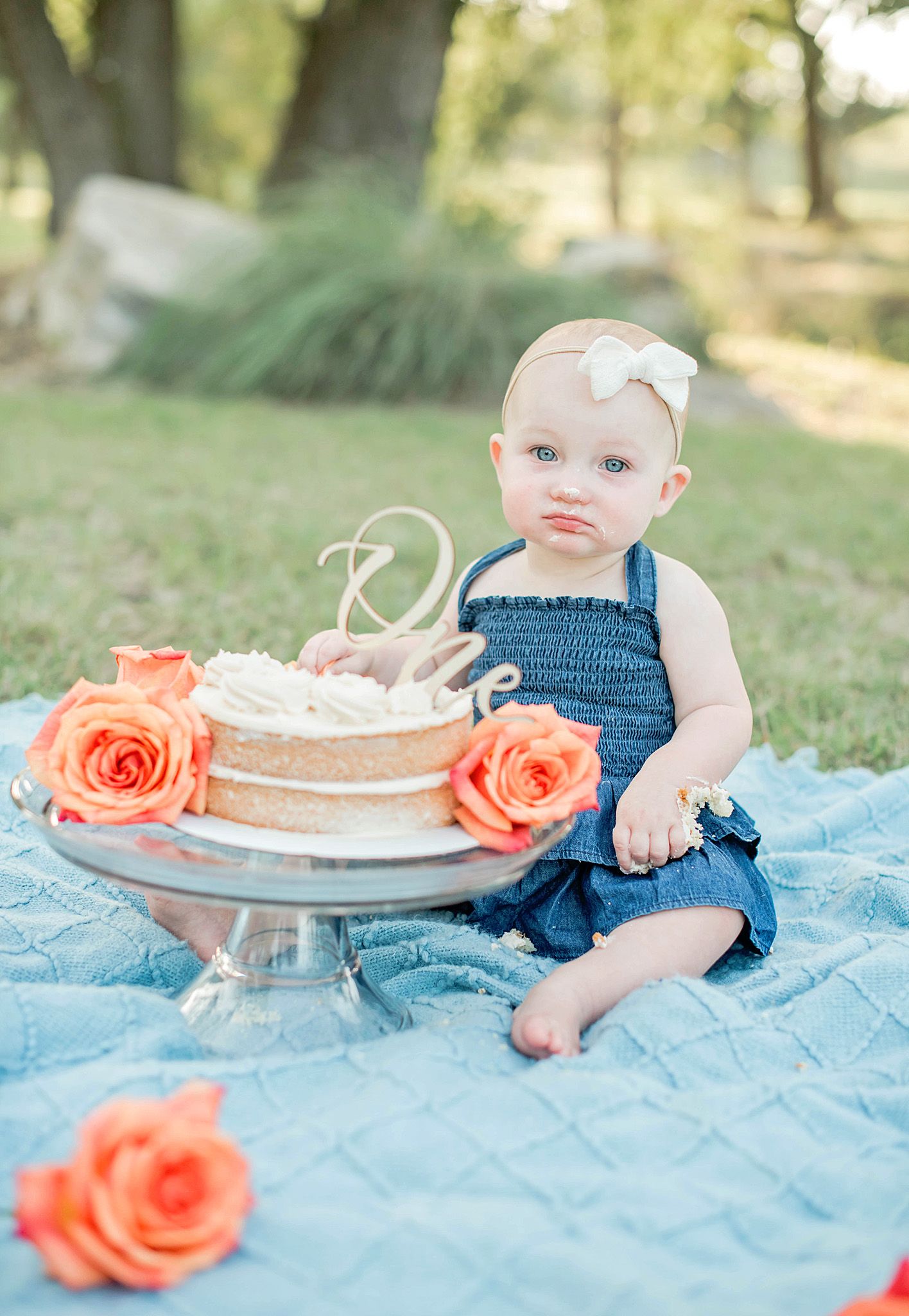 Baby girl in denim romper sits beside a decorated first birthday cake with orange roses on a blue blanket outdoors.