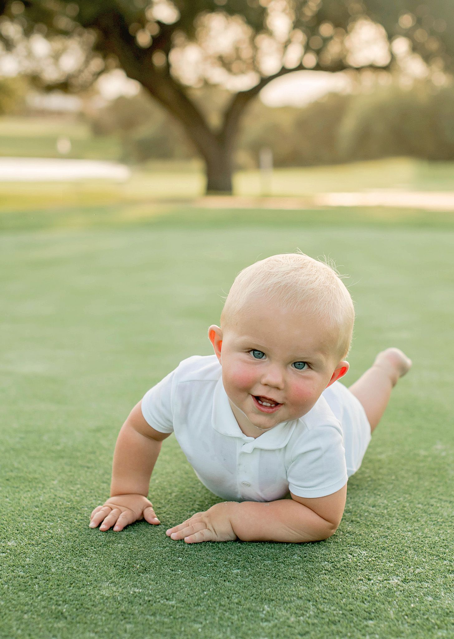 Sweet baby in white onesie crawls and smiles on lush green grass during an outdoor photo session.